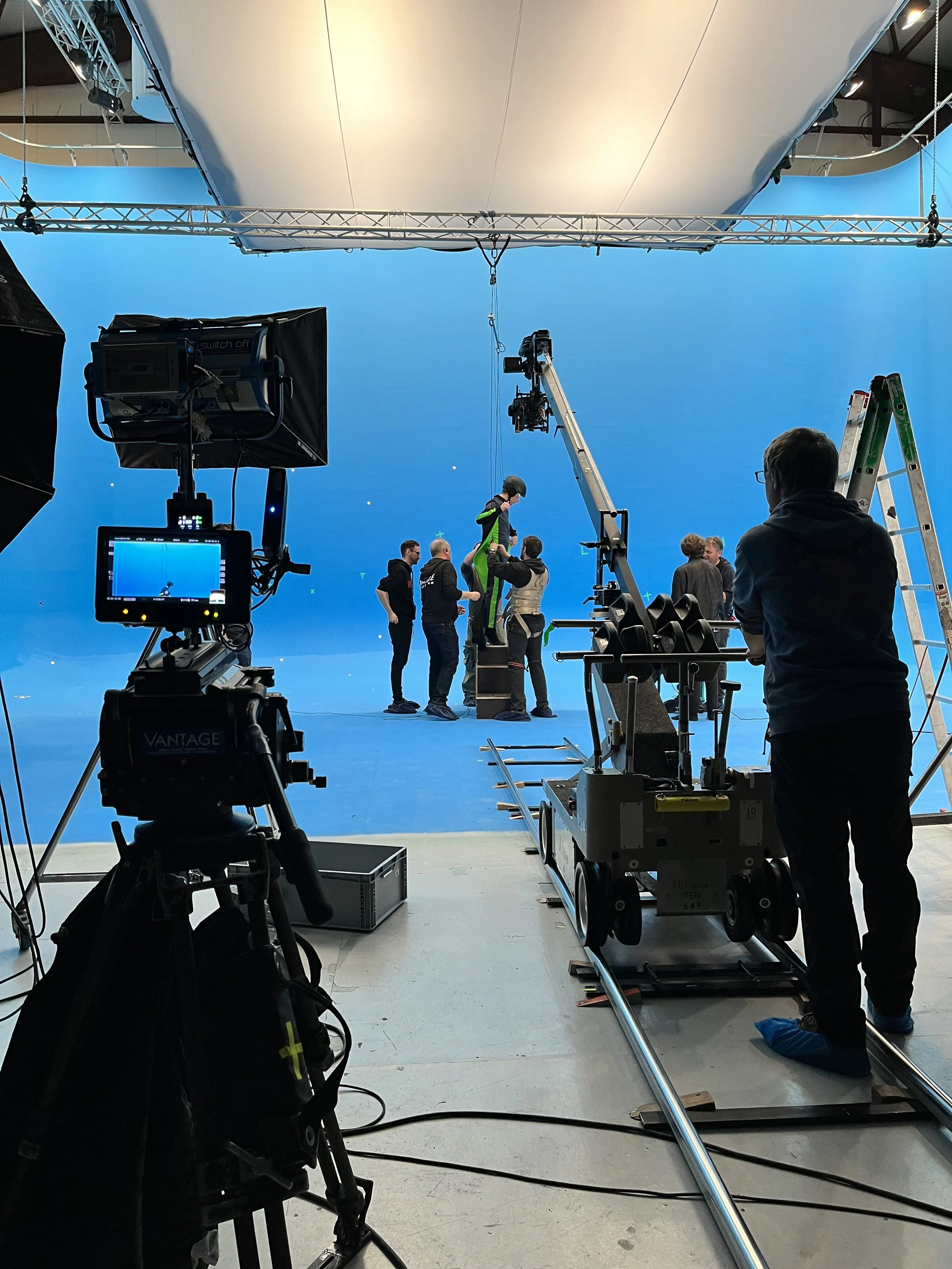 Film crew working on a production shoot with a blue screen backdrop, lighting equipment, and cameras in a studio with crew members adjusting gear and actors preparing for a scene.