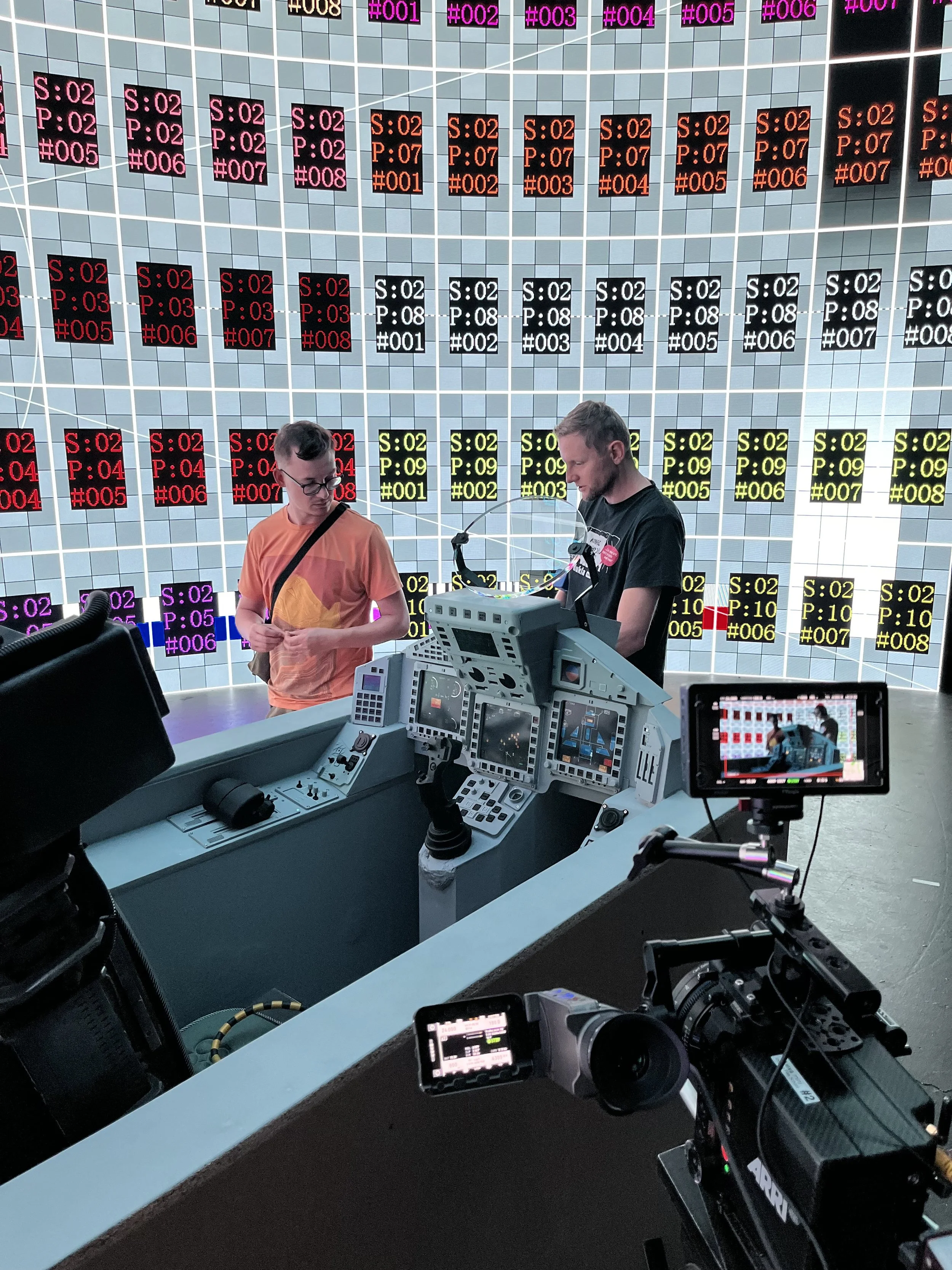 Inside an airport control or operations center with multiple digital display screens showing flight information, and two staff members working at a console surrounded by cameras and equipment.