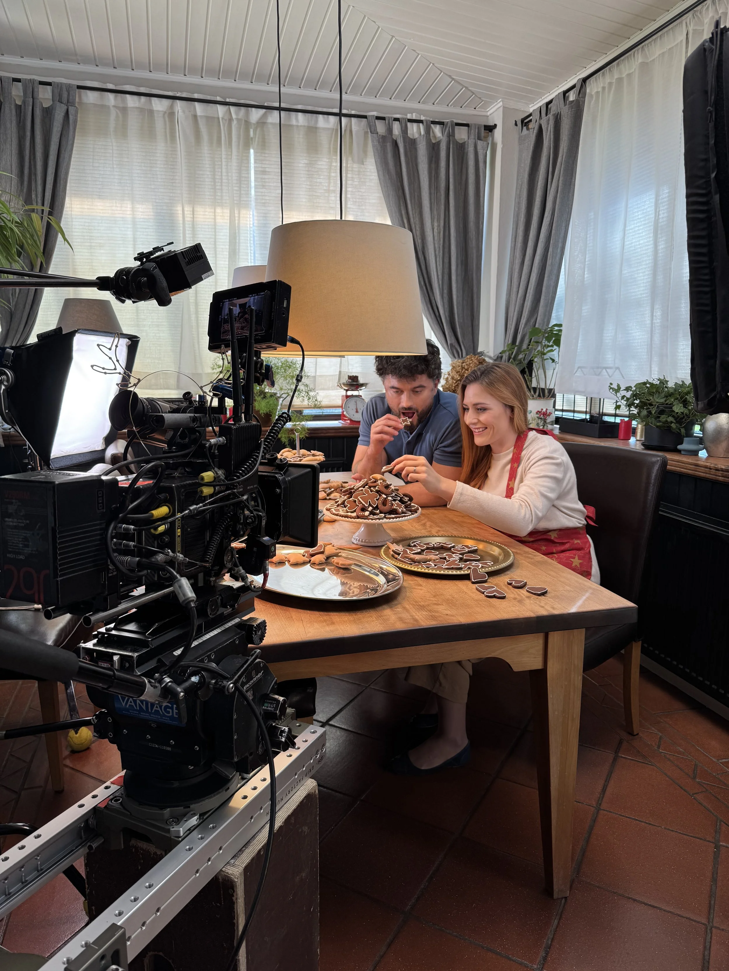 Two people sitting at a wooden table, decorating cookies with gingerbread men and other shapes, with a camera and filming equipment set up nearby, in a cozy room with curtains and plants.