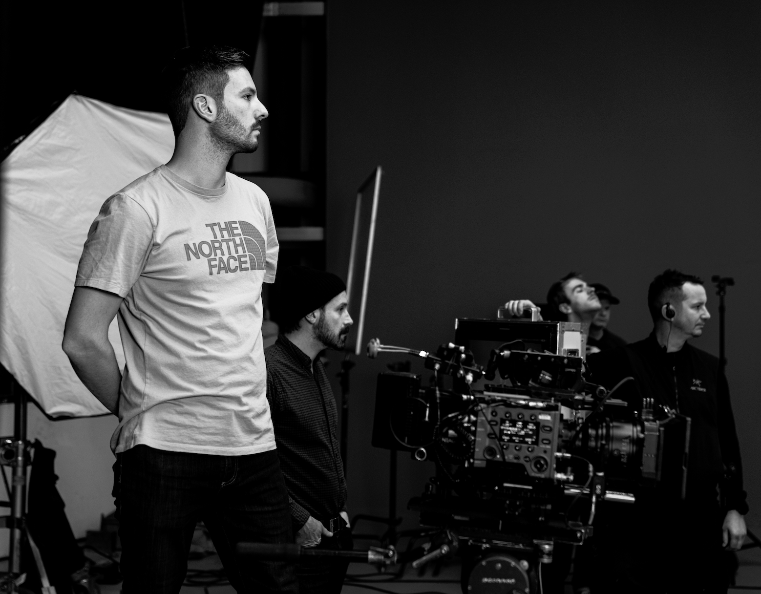 A man wearing a North Face t-shirt stands with his hands behind his back, looking to the left, on a film set with camera crew members and equipment.