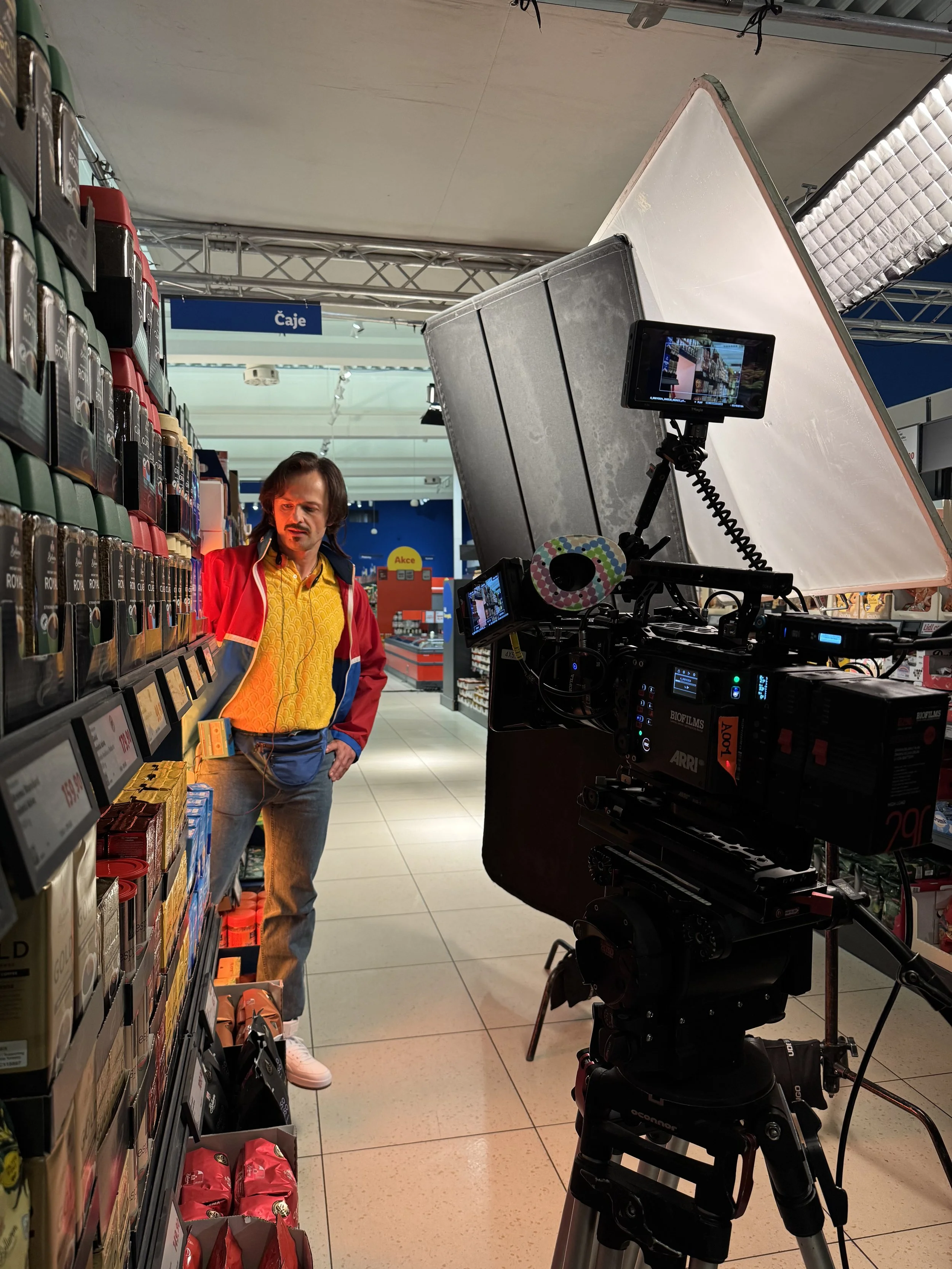 A man with long dark hair, wearing a colorful jacket and yellow shirt, is standing in a grocery store aisle, looking at the products on the shelf. A professional camera and lighting equipment are set up nearby, indicating a filming setup inside the store.