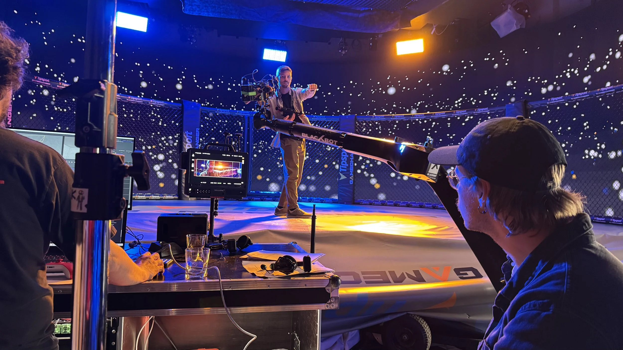 A man operating a camera on a stabilizer in a staged cage fight environment with blue galaxy-themed lighting and an audience watching.