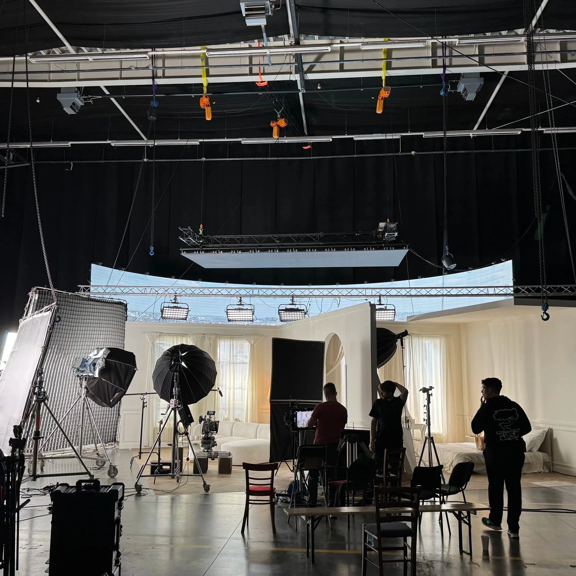 Inside a studio with professional lighting and camera crew preparing for filming, with a white sofa, large windows with curtains, and a black backdrop.