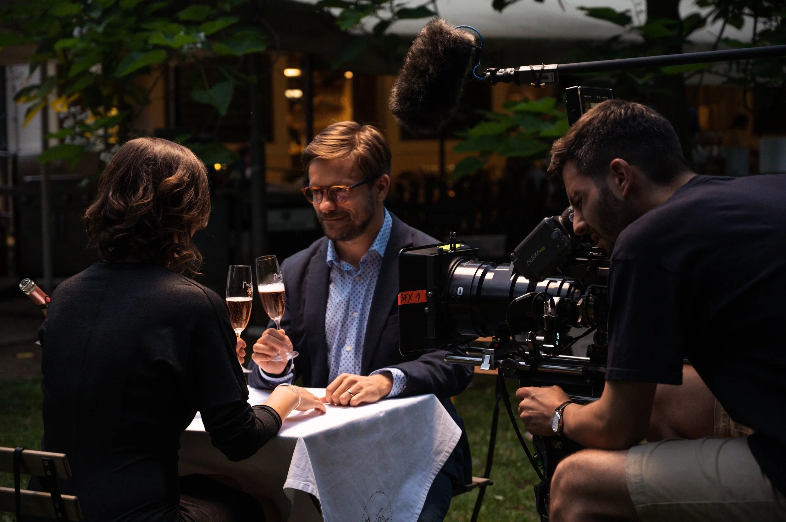 Filming a romantic scene at an outdoor wedding with a camera crew, a couple holding glasses of champagne, and a person operating a camera in the foreground.