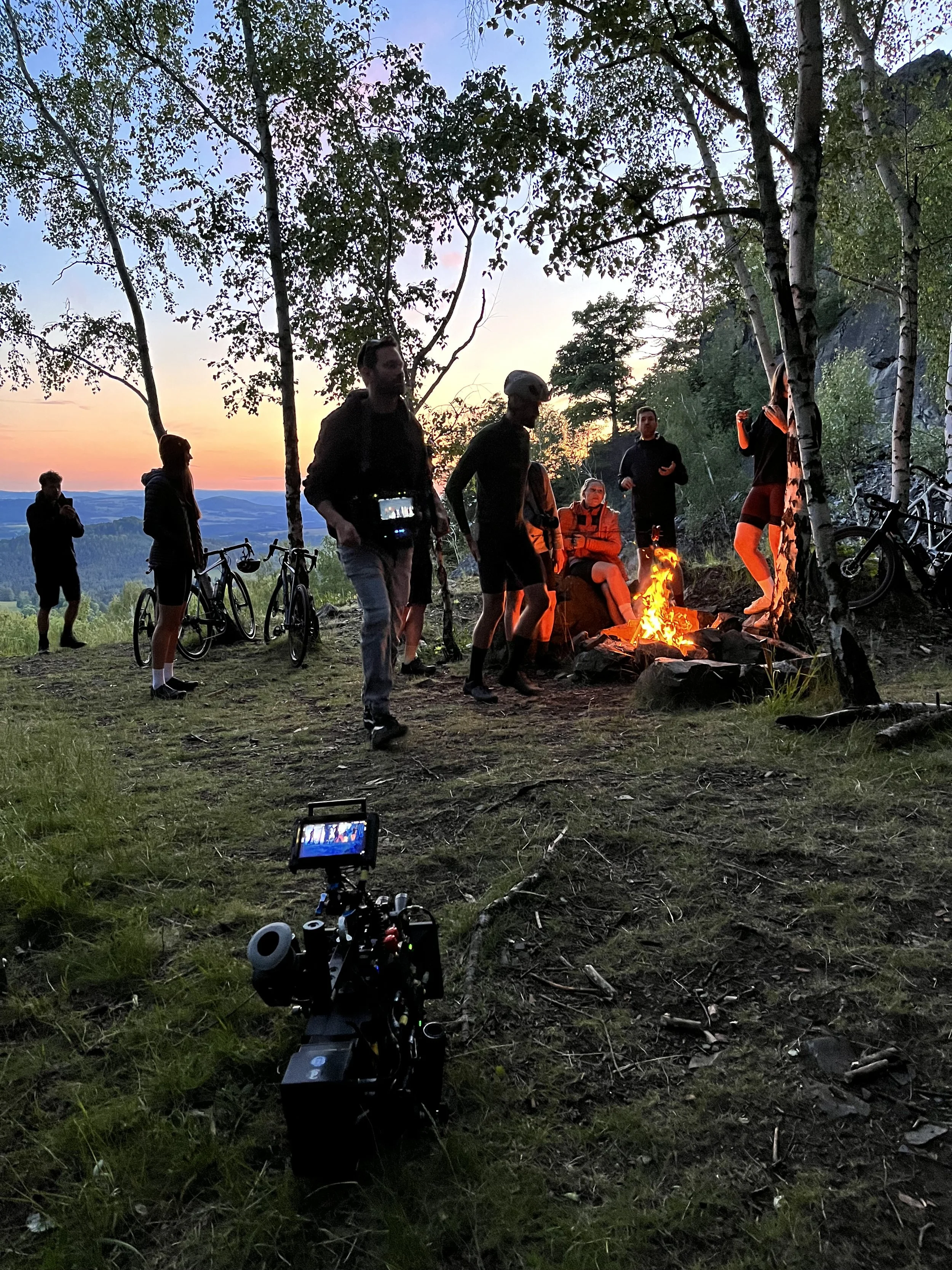 Group of people gathered around a campfire in a forest during sunset, with bicycles nearby and camera equipment on the ground.