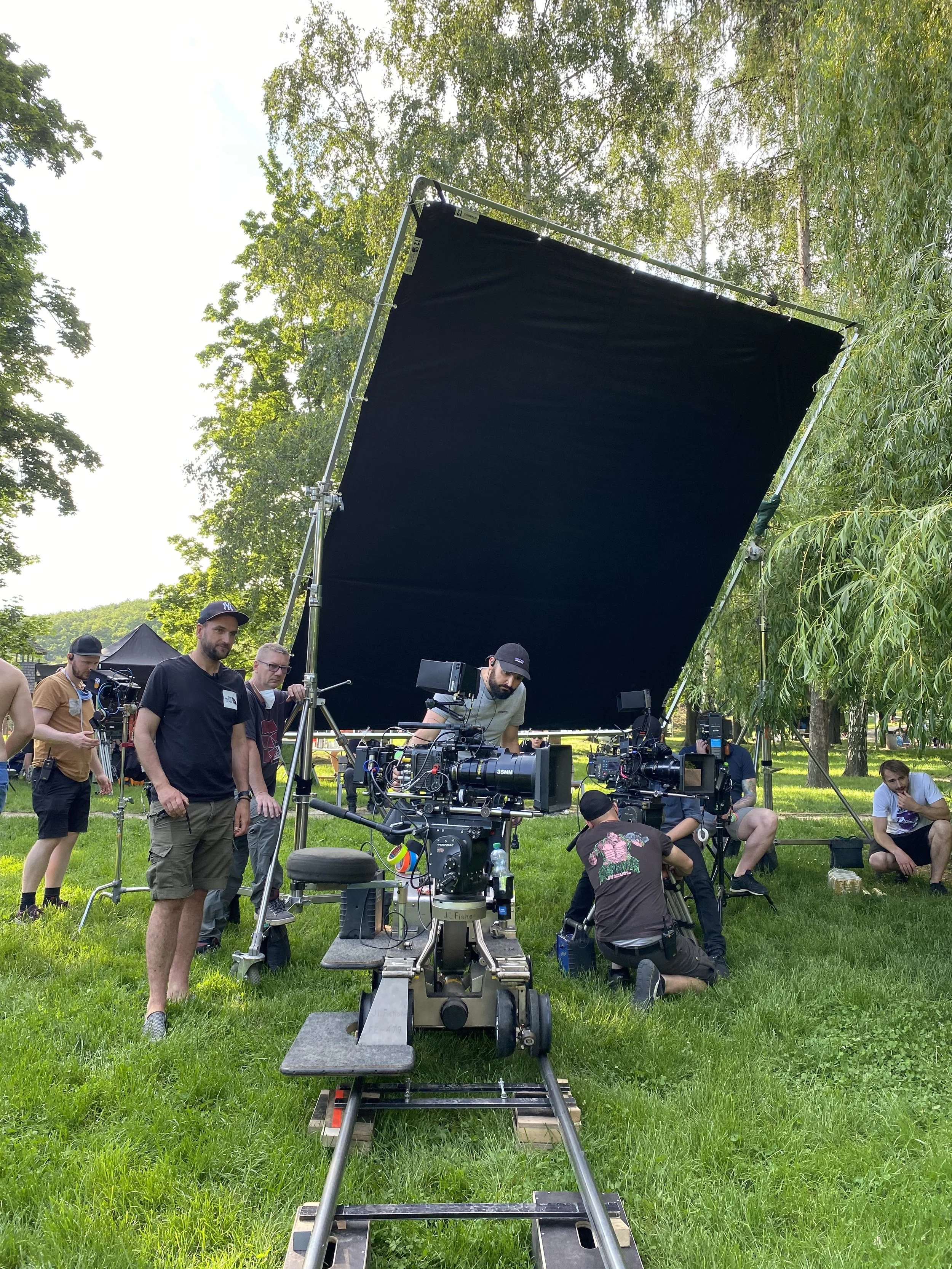A film crew working outdoors on a filming set, with a large black screen and cameras positioned on a track, surrounded by green trees and grass.