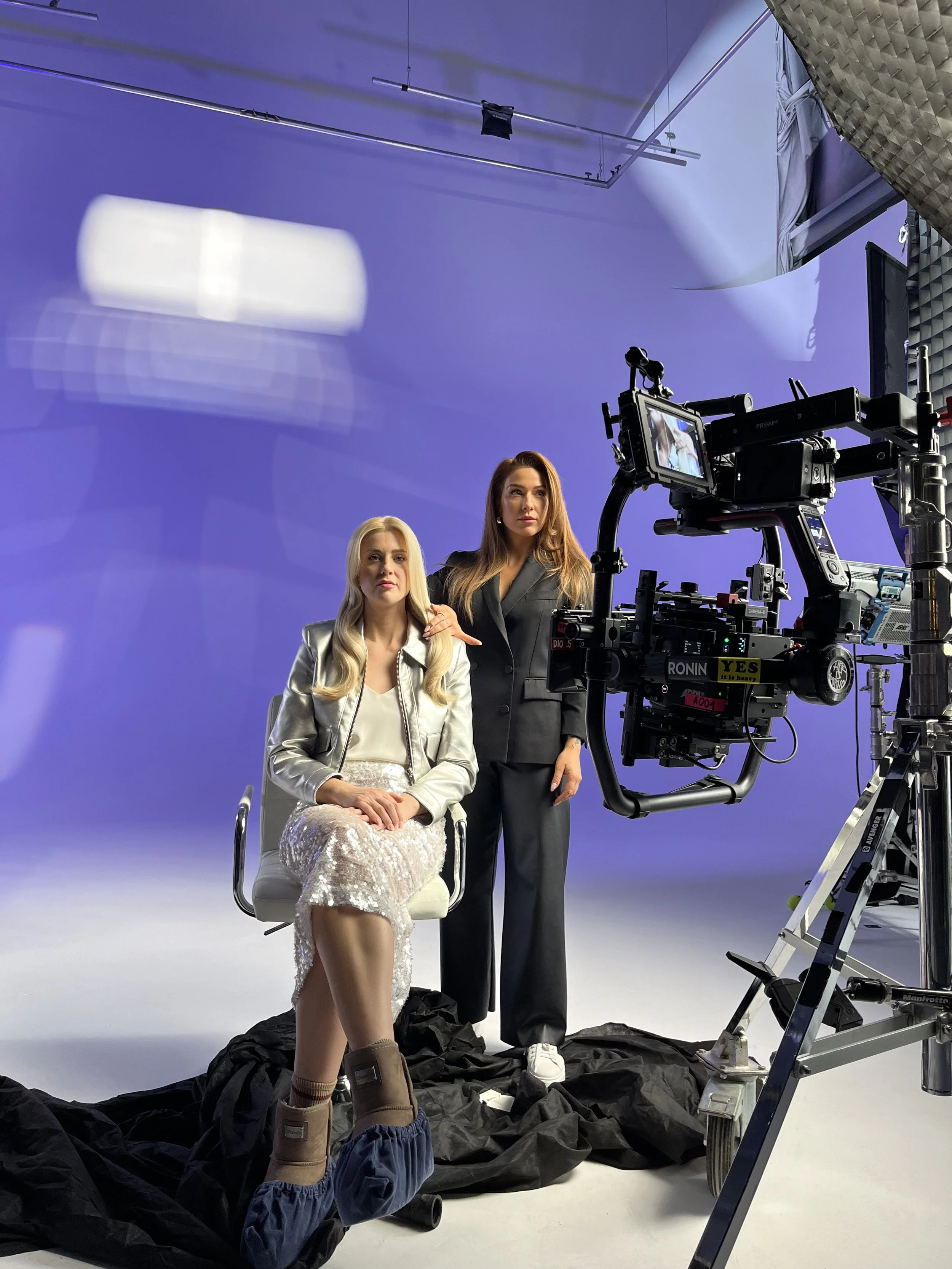Two women on a photoshoot set, one sitting wearing a shiny white dress and the other standing with one hand on the seated woman's shoulder, against a purple studio background, with professional camera equipment and crew members visible.