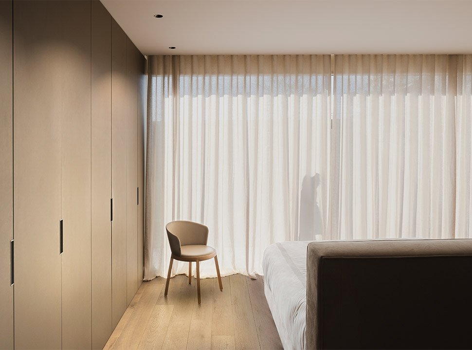 En moderne hotelværelse med en seng, en beige stol, store gardiner og indbyggede skabe på væggen.