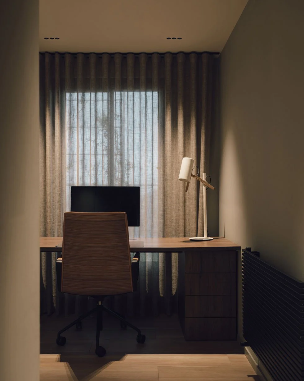Et minimalistisk hjemmekontor med et skrivebord, en kontorstol, en computerskærm, en hvid skrivebordslampe og lange gardiner foran et vindue.