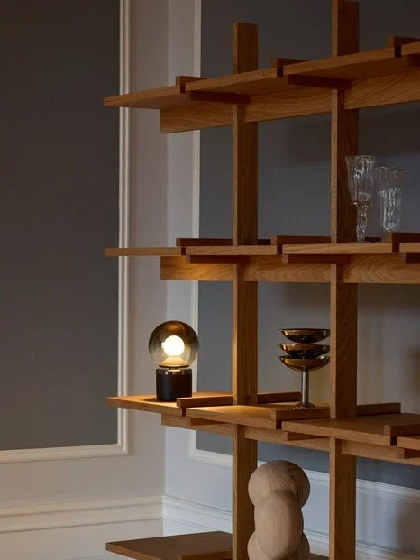 En moderne træreol med tre hylder, der indeholder et glas, nogle små fade, en lampe med en globus-kugle, og en træfigur, placeret i et minimalistisk hjem med neutral vægfarve.