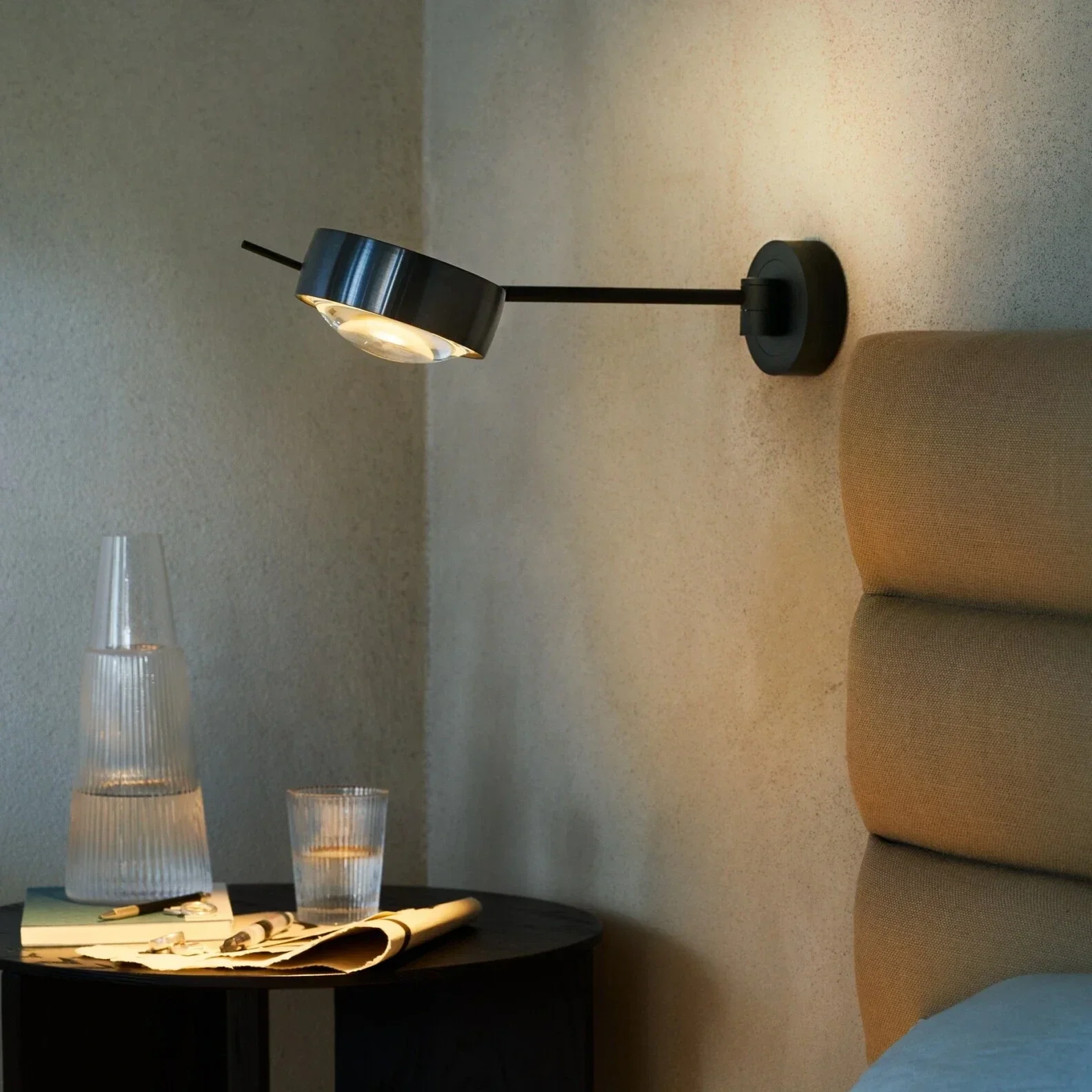 En sort rund bord med et glasvase, to vinglas, en avis, nogle nøgler og en lighter. En væg med en væglampe og en beige sofa.