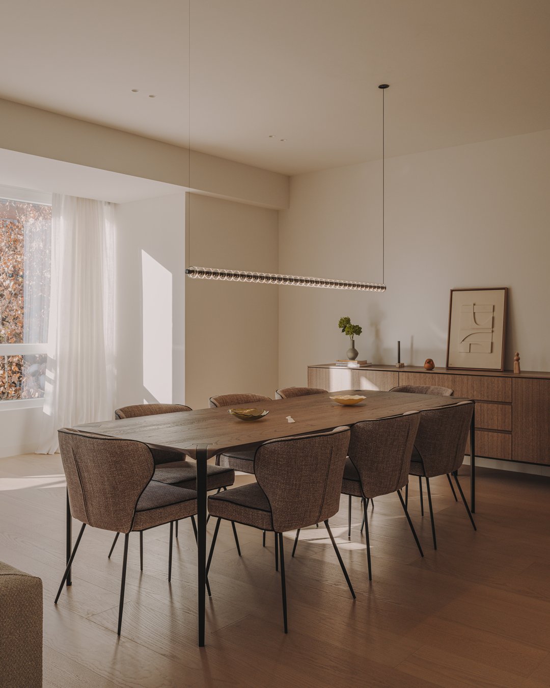 Moderne spisestue med træbord, otte polstrede stole, stor vindue med hvide gardiner, minimalistisk vægdekoration og et skab med dekorative genstande.