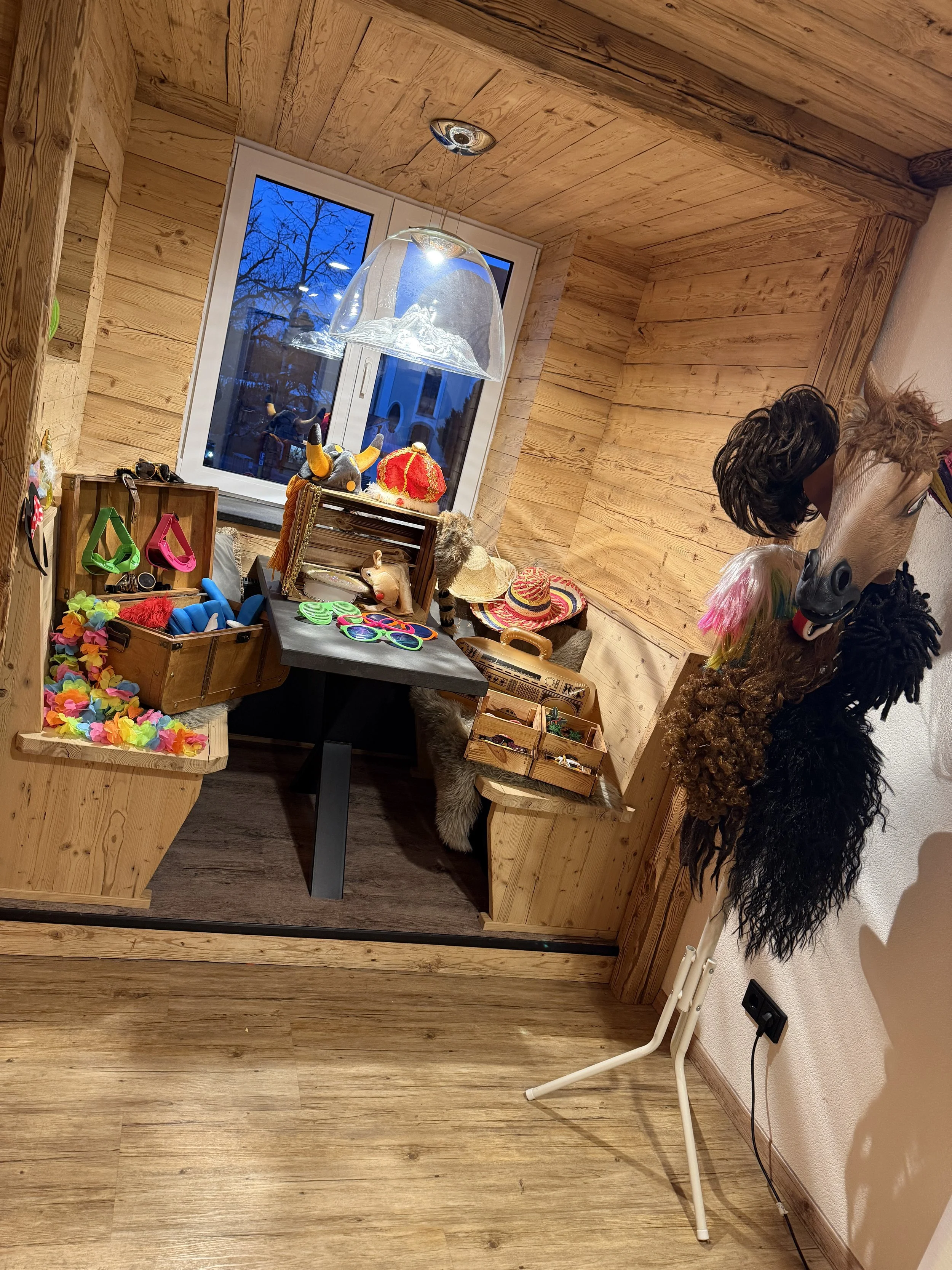 Ein kleines Zimmer mit Holzverkleidung, dekoriert mit verschiedenen lustigen Kopfbedeckungen und Accessoires, wahrscheinlich eine Verkleidungsstation, mit einem Fenster, das nach draußen in eine winterliche Landschaft blickt.