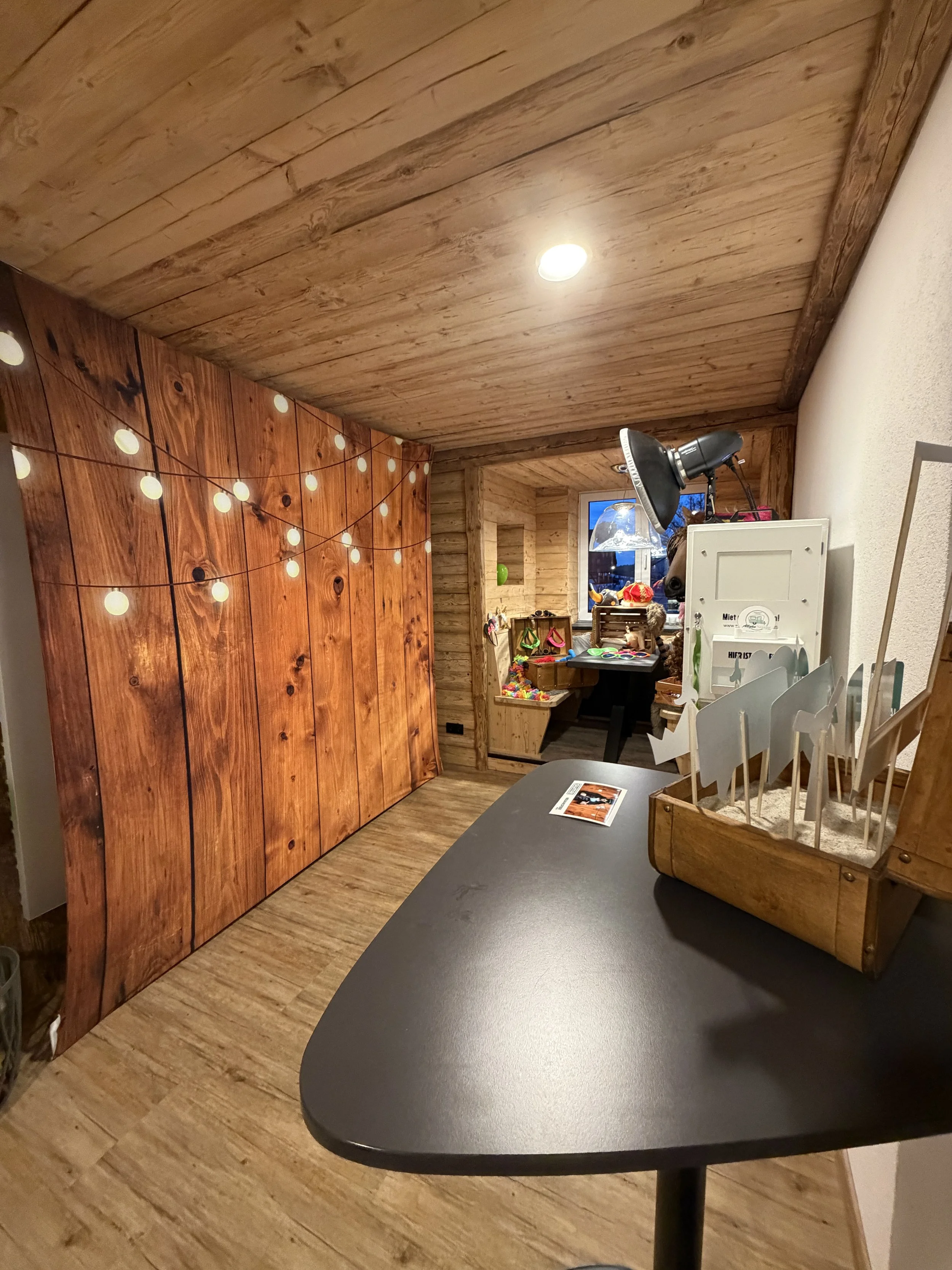 Ein Raum mit Holzverkleidung an den Wänden und Decke, eine Tischplatte mit einem Flyer, eine Holzkiste mit Briefen, eine Servicestation mit Lampen, Bastelmaterialien und Dekorationen im Hintergrund, verbunden mit einer gemütlichen Atmosphäre.