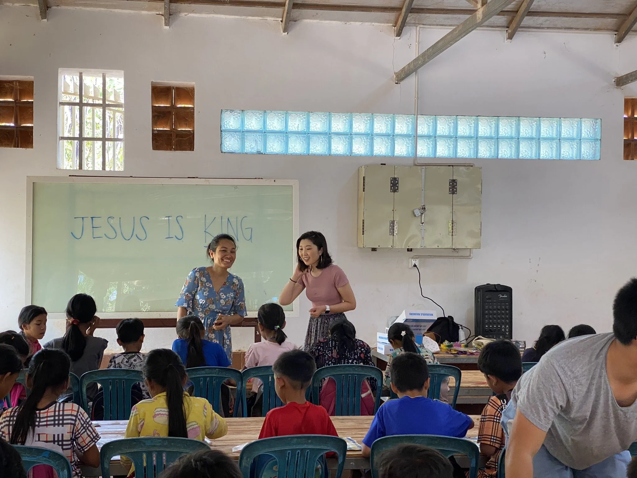 Classroom setting with a message "JESUS IS KING" on the whiteboard, two women standing in front of students seated at tables.