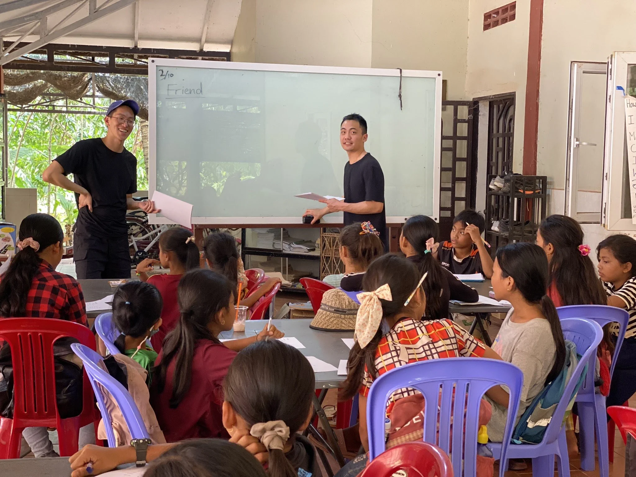 A classroom with two instructors standing at the front near a whiteboard, teaching a group of students sitting on colorful plastic chairs.