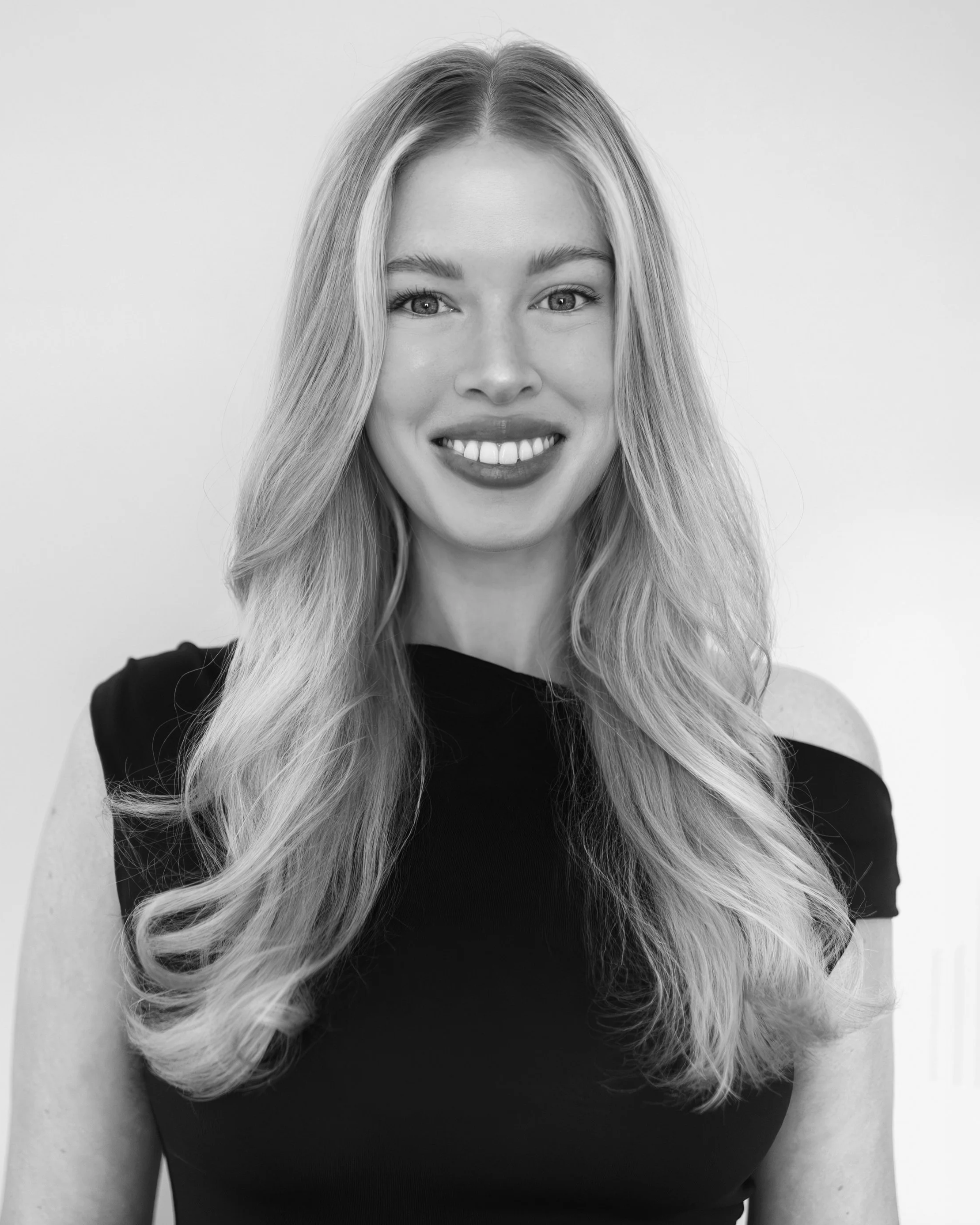 Black and white portrait of a woman with long wavy hair, smiling, wearing a cold shoulder top, and standing against a plain background.