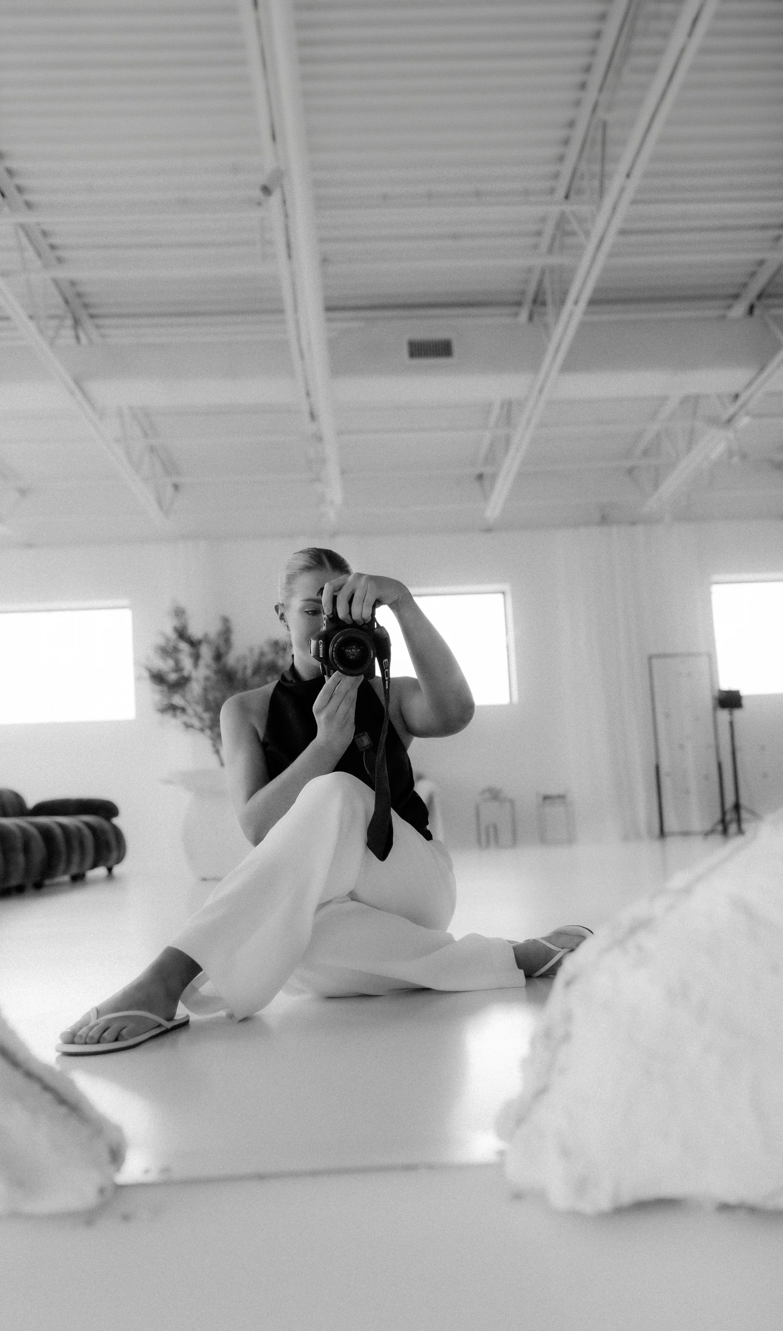 A woman taking a photo of herself with a camera in a bright room with large windows, a high ceiling, and minimal decor.