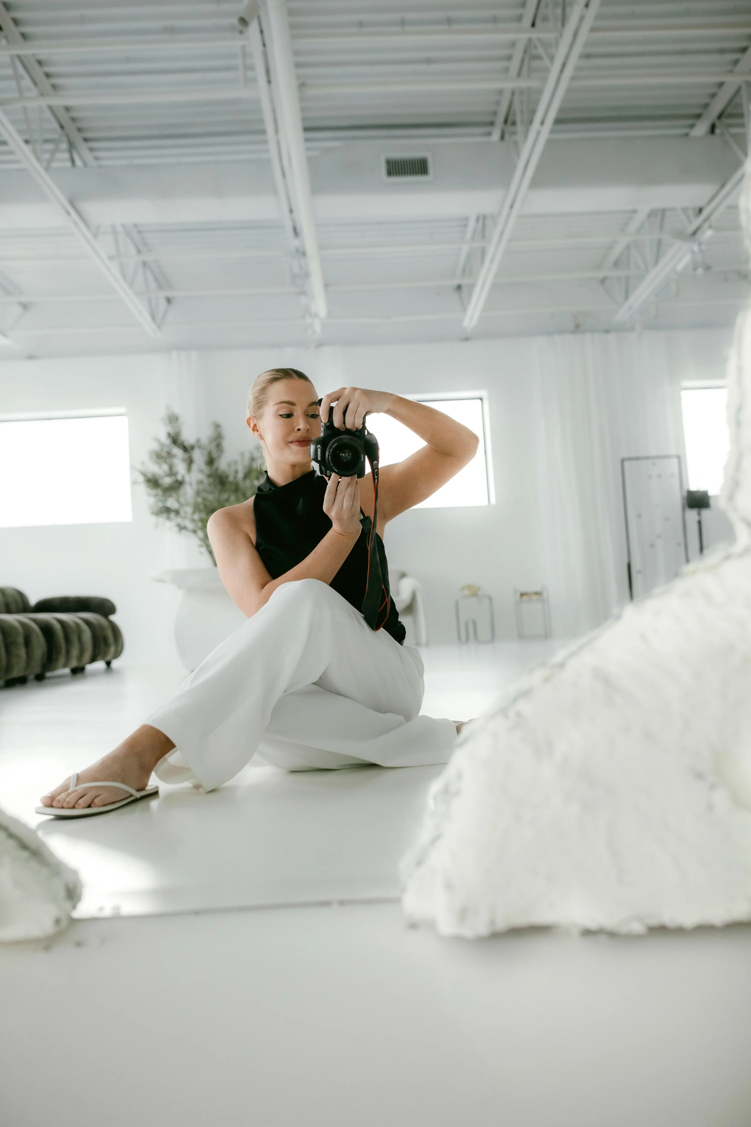 A woman sitting on the floor in a bright, minimalist room, taking a selfie with a camera. She is wearing a black sleeveless top, white pants, and white sandals, with her hair tied back. The room has large windows, white walls, and some furniture and greenery in the background.