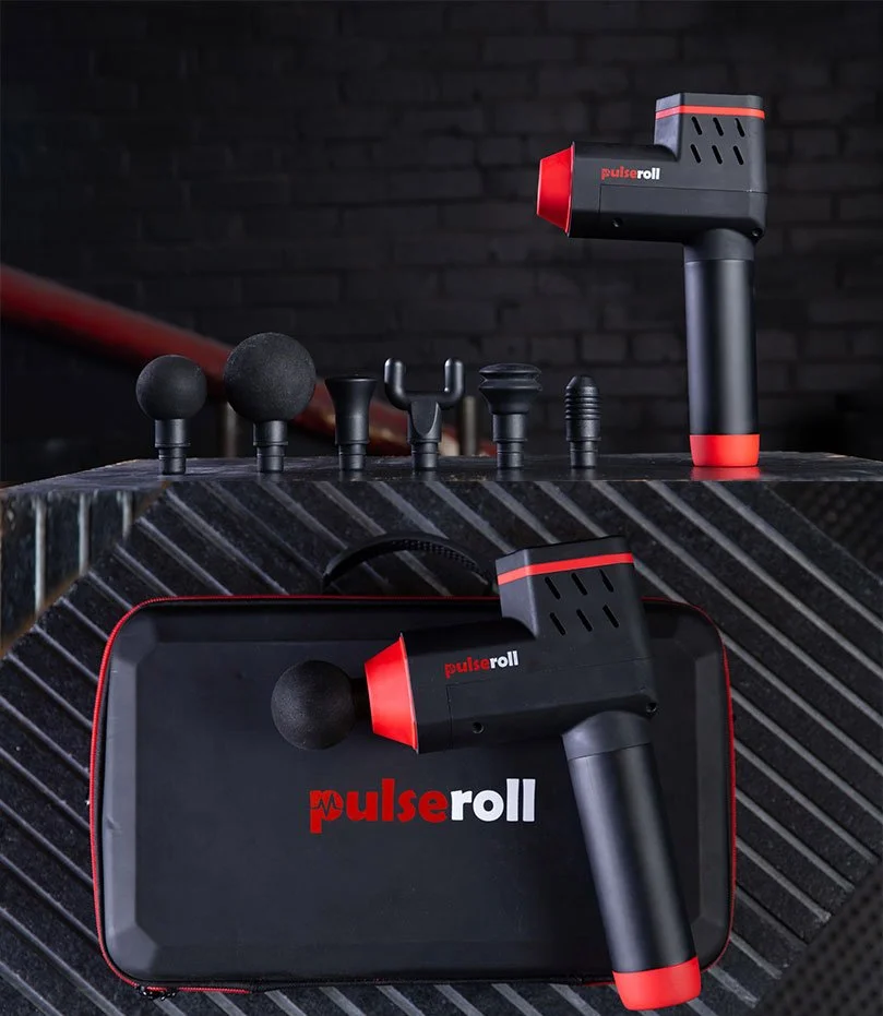 A black and red percussion massage gun with several interchangeable heads and carrying case on a textured surface, with additional massage heads in the background against a dark brick wall.