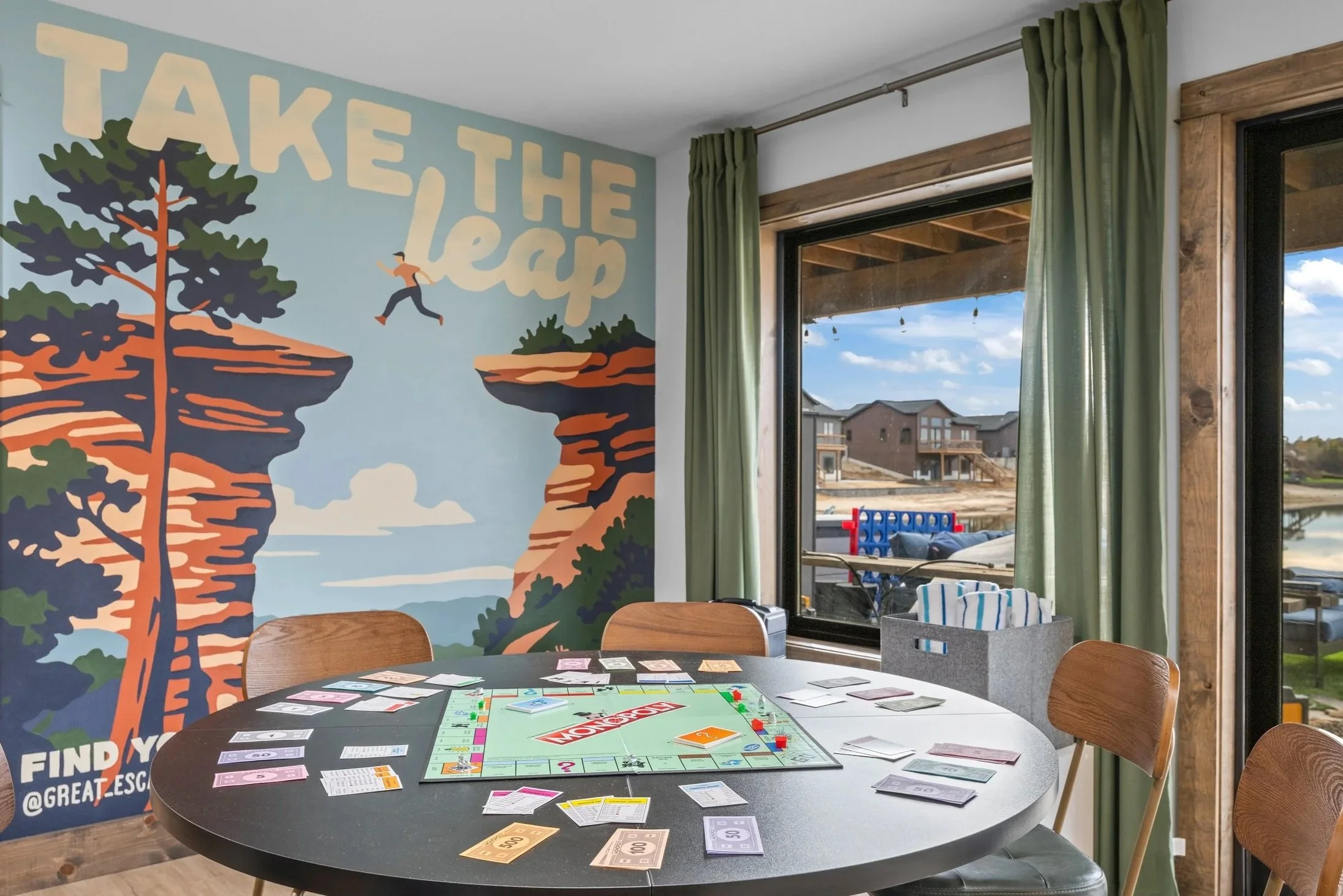 Person kneeling on floor, stirring white paint in a large paint can with a wooden stir stick. There are other paint supplies around, including additional paint cans, a paintbrush, and scissors. The person is wearing a green jacket covered in white paint splatters and light gray sweatpants.