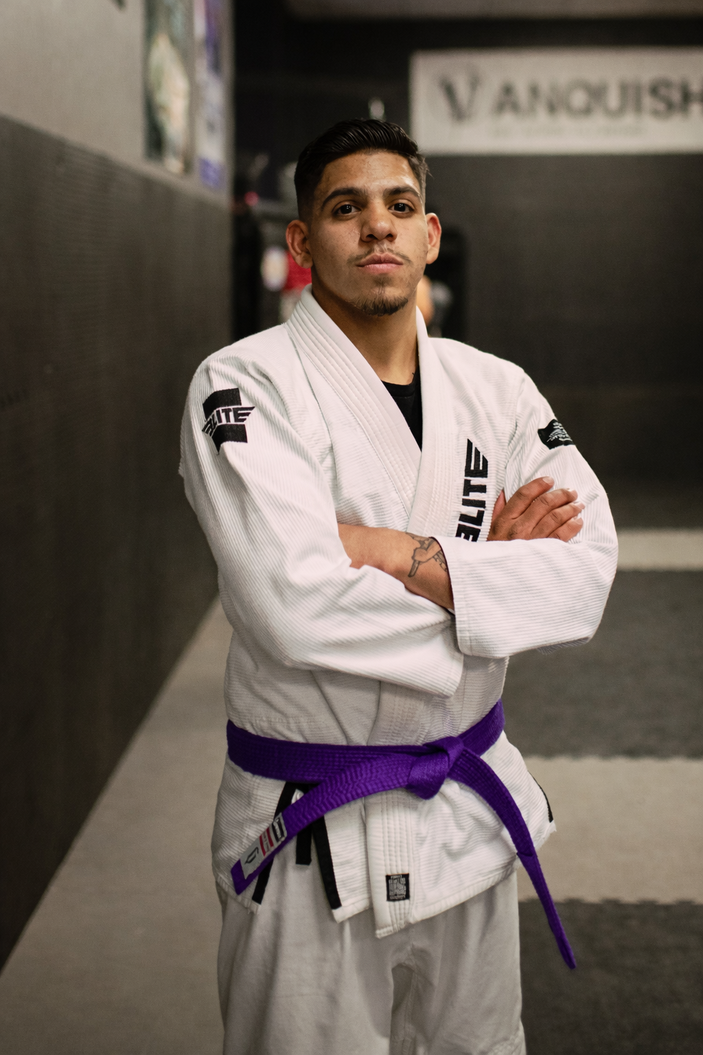 This is an image of a jujitsu practitioner standing in a gym wearing a white guy with a purple belt with his arms crossed