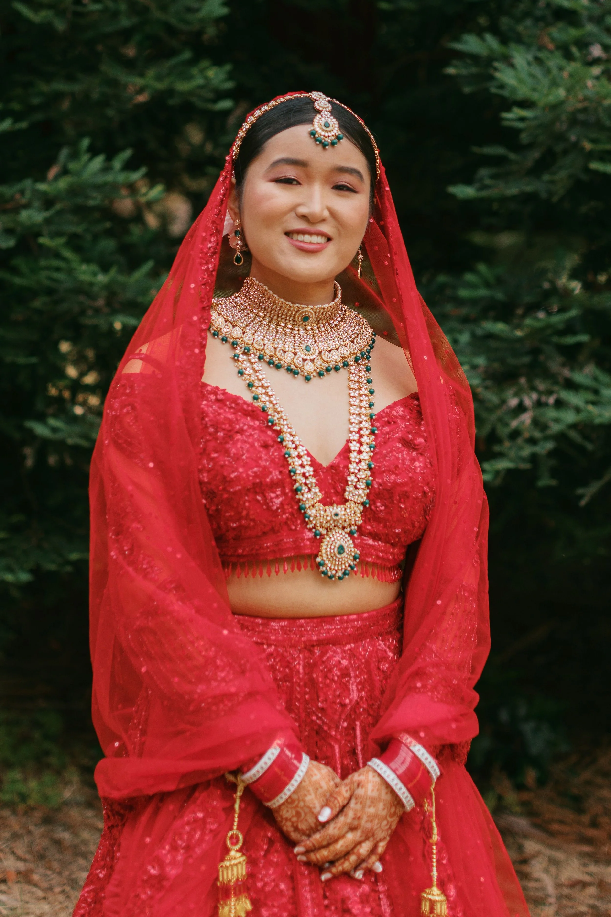 A woman dressed in traditional South Asian bridal attire with red and gold jewelry, necklaces, earrings, and henna on her hands, standing outdoors in front of green foliage.