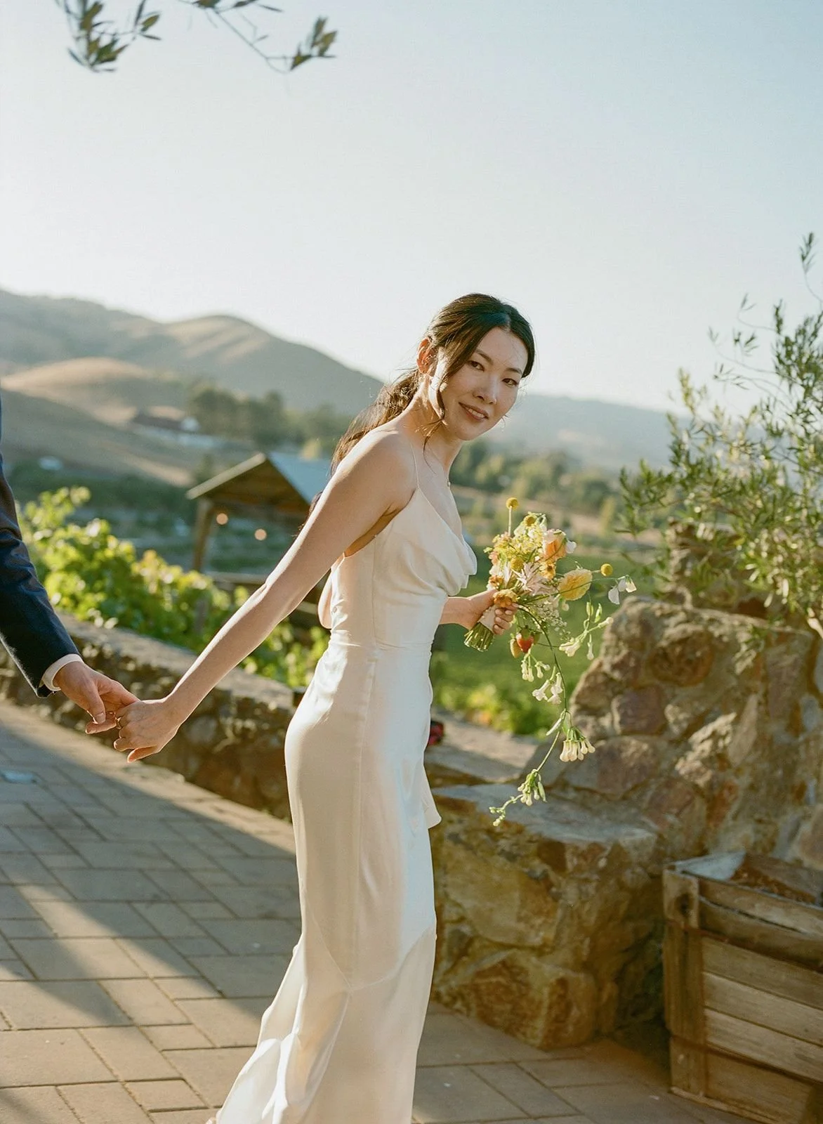 A woman in a white dress holding a bouquet of flowers, standing outdoors on a paved path with mountains and greenery in the background.