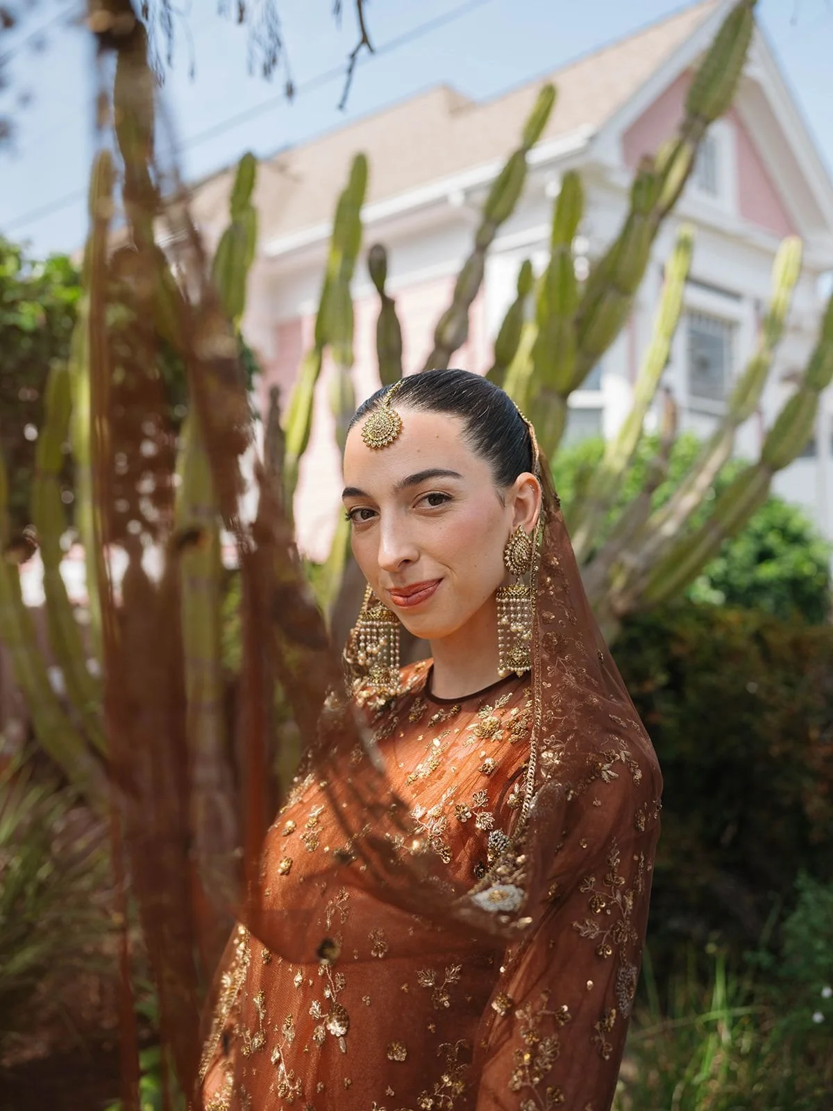A woman in traditional Indian attire with jewelry, posing outdoors with a cactus plant and a pink house in the background.