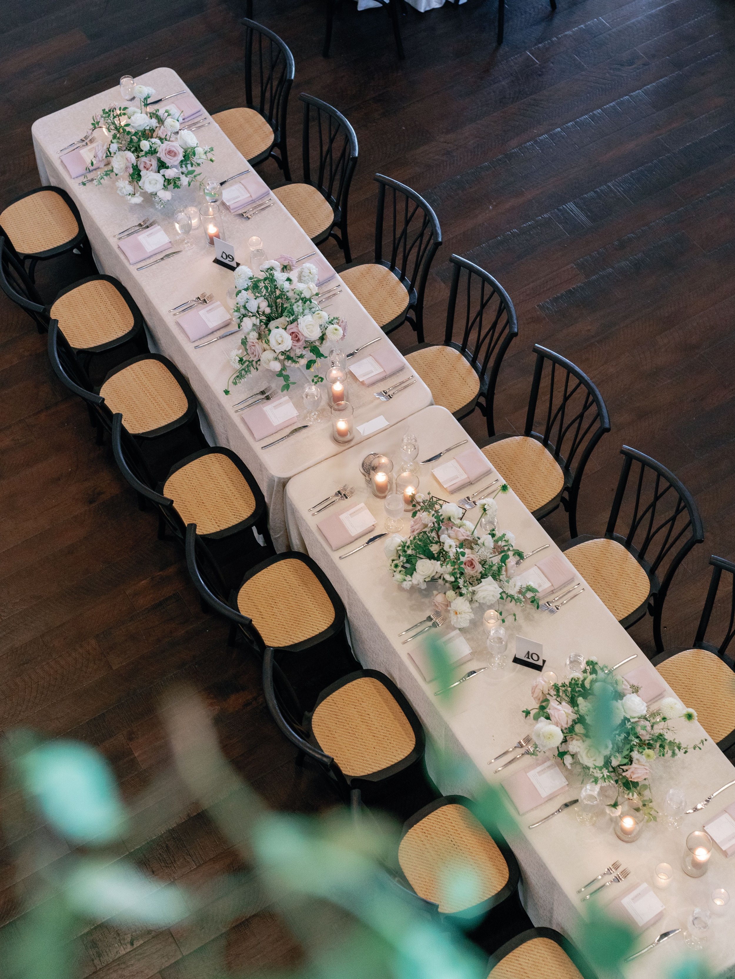 A long banquet table decorated with white and pink floral centerpieces, candles, pink napkins, and silverware in a dimly lit venue with dark wooden floors.