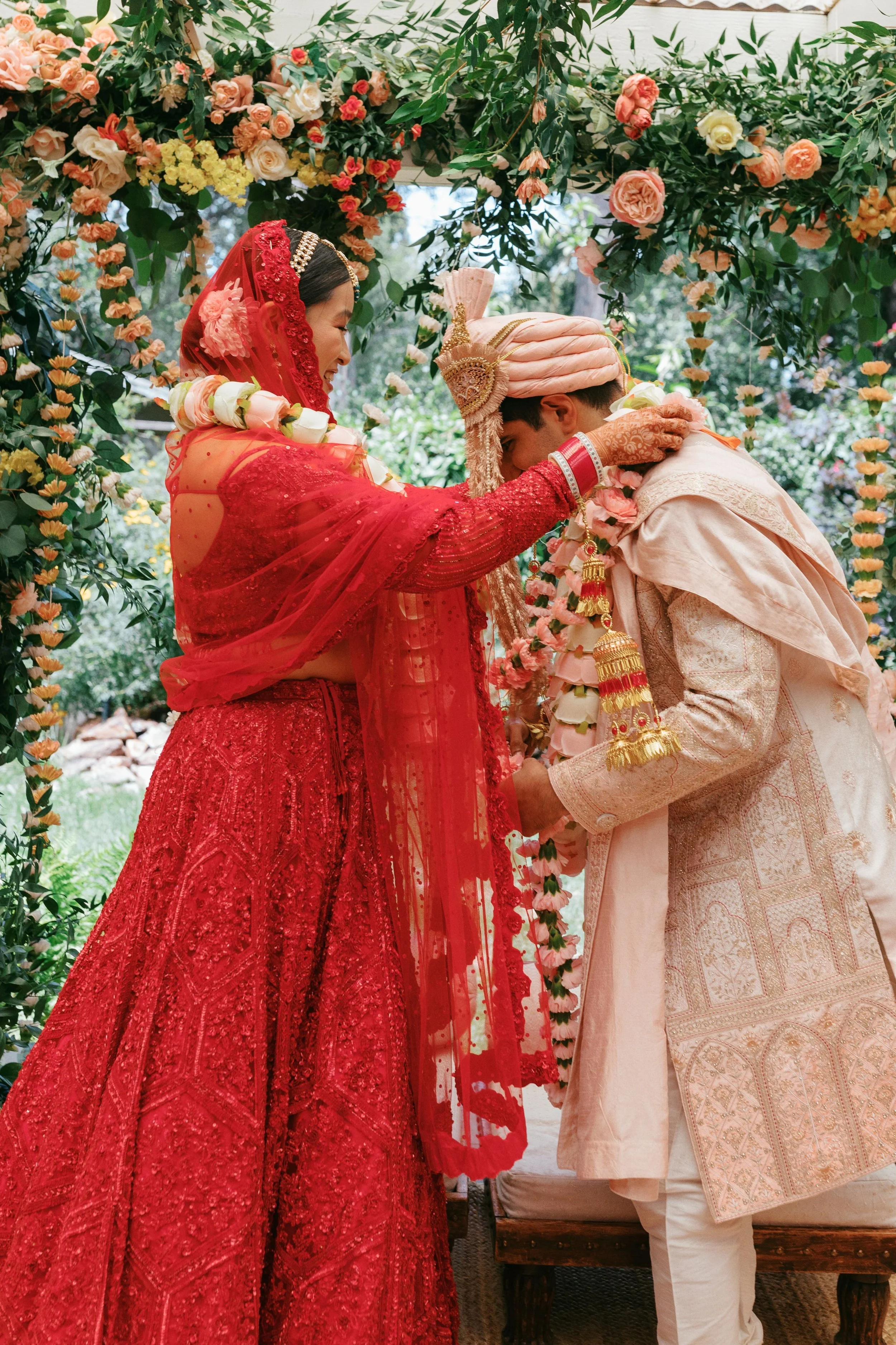 A bride and groom at their wedding ceremony, dressed in traditional Indian attire, exchanging garlands under floral decorations.