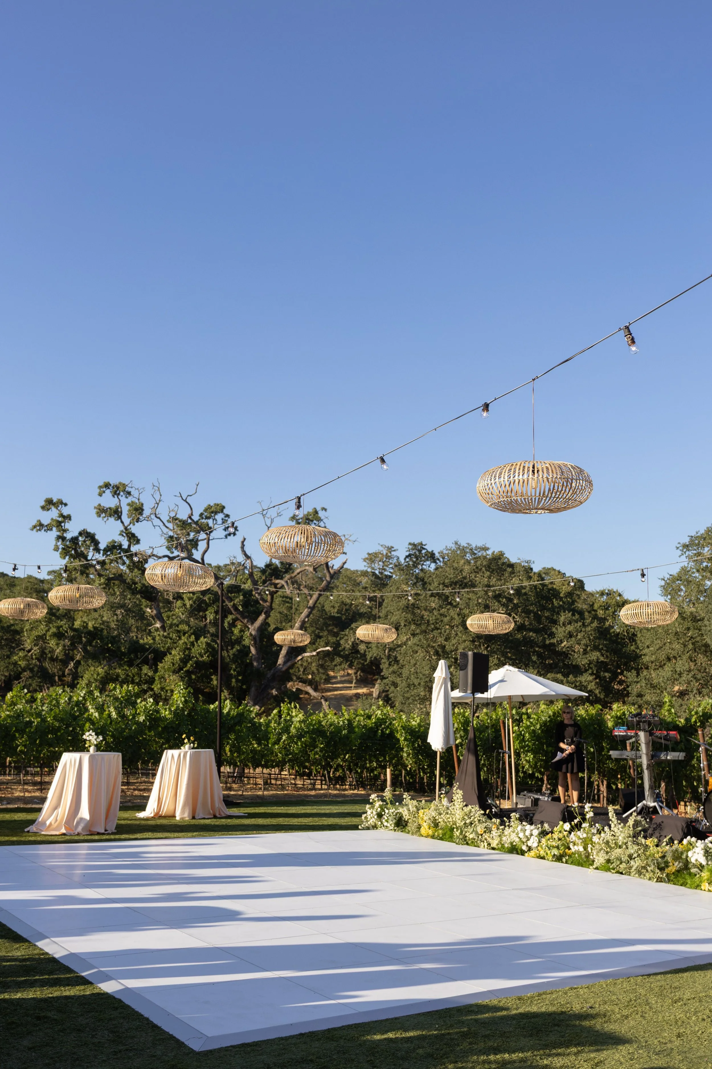 An outdoor wedding reception setup with a white dance floor, decorative hanging lanterns, tables with beige tablecloths, white and yellow flowers, and a musician preparing to perform. There are trees and vineyard in the background with string lights 