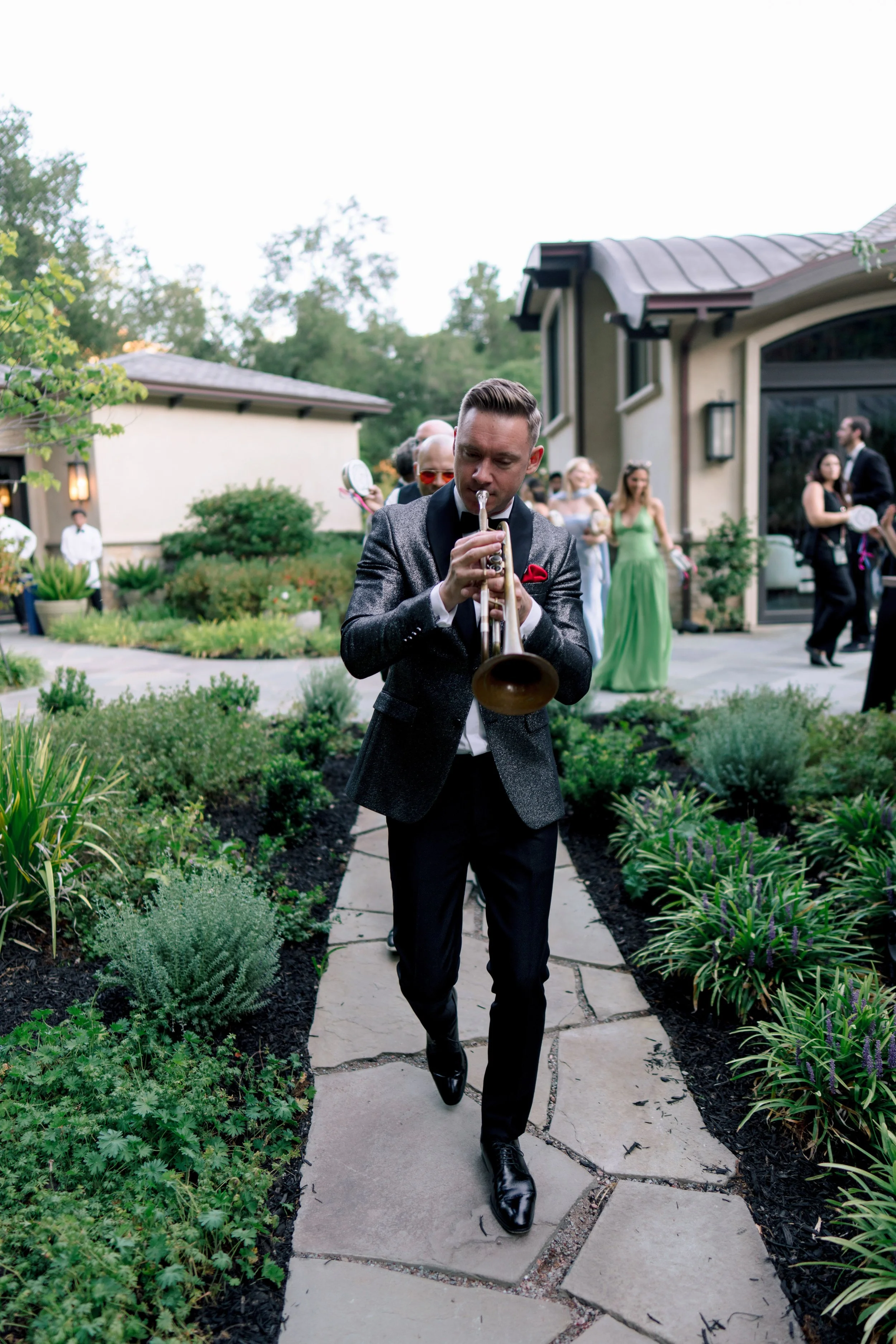 A man in a tuxedo shirt and black shoes walking on a garden pathway while playing a trumpet at an outdoor event. Several people are in the background, some holding plates, and there is a house with large windows and a metal roof.