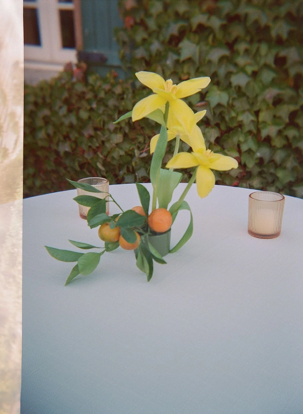 A flower arrangement with yellow orchids and green leaves in a small black pot on a white table, with two small glass candle holders nearby. In the background, there is a hedge and parts of a house exterior.