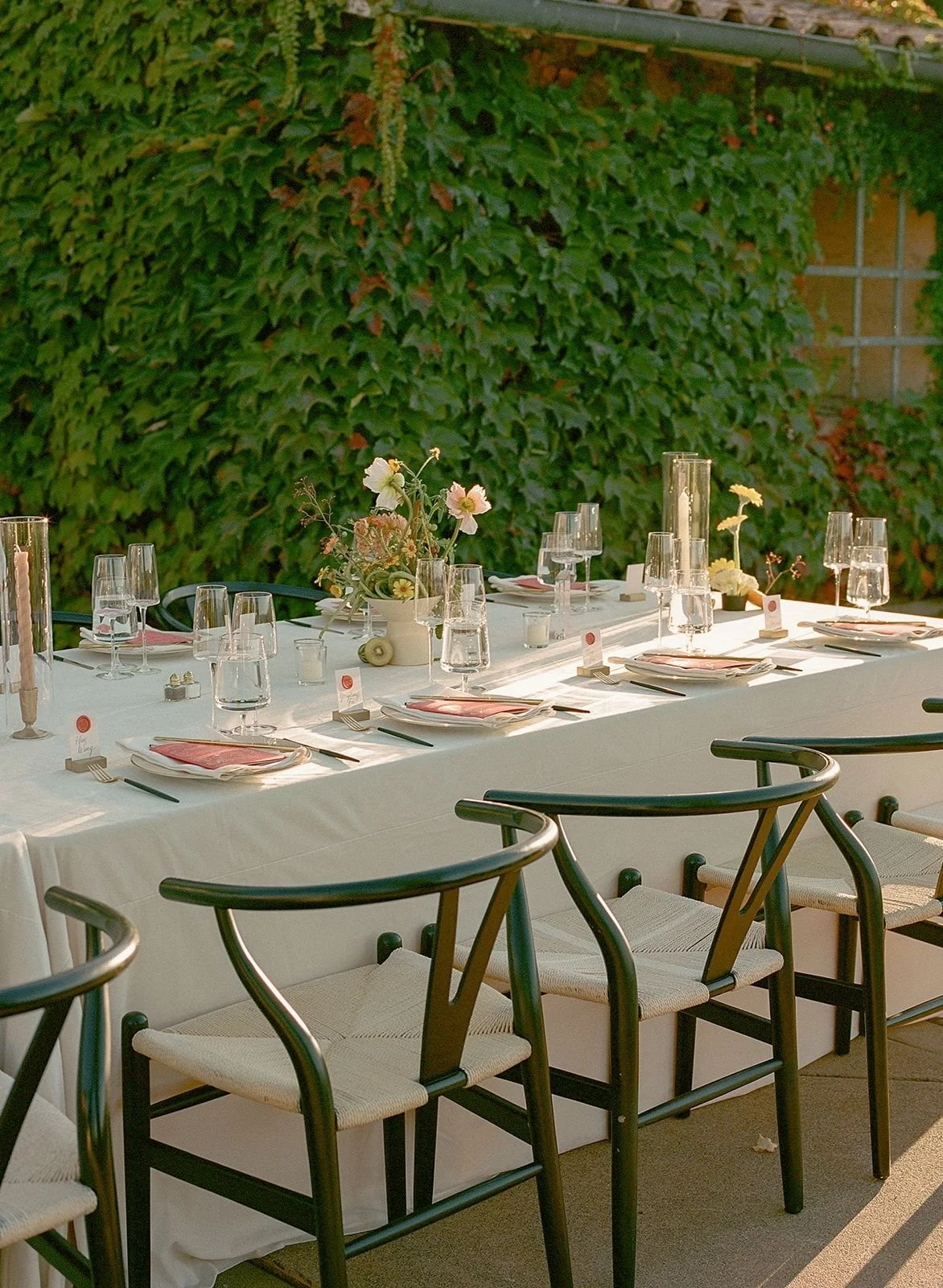 Outdoor dining table set with white tablecloth, pink napkins, glassware, flower arrangements, and candles, against a background of green ivy-covered wall.