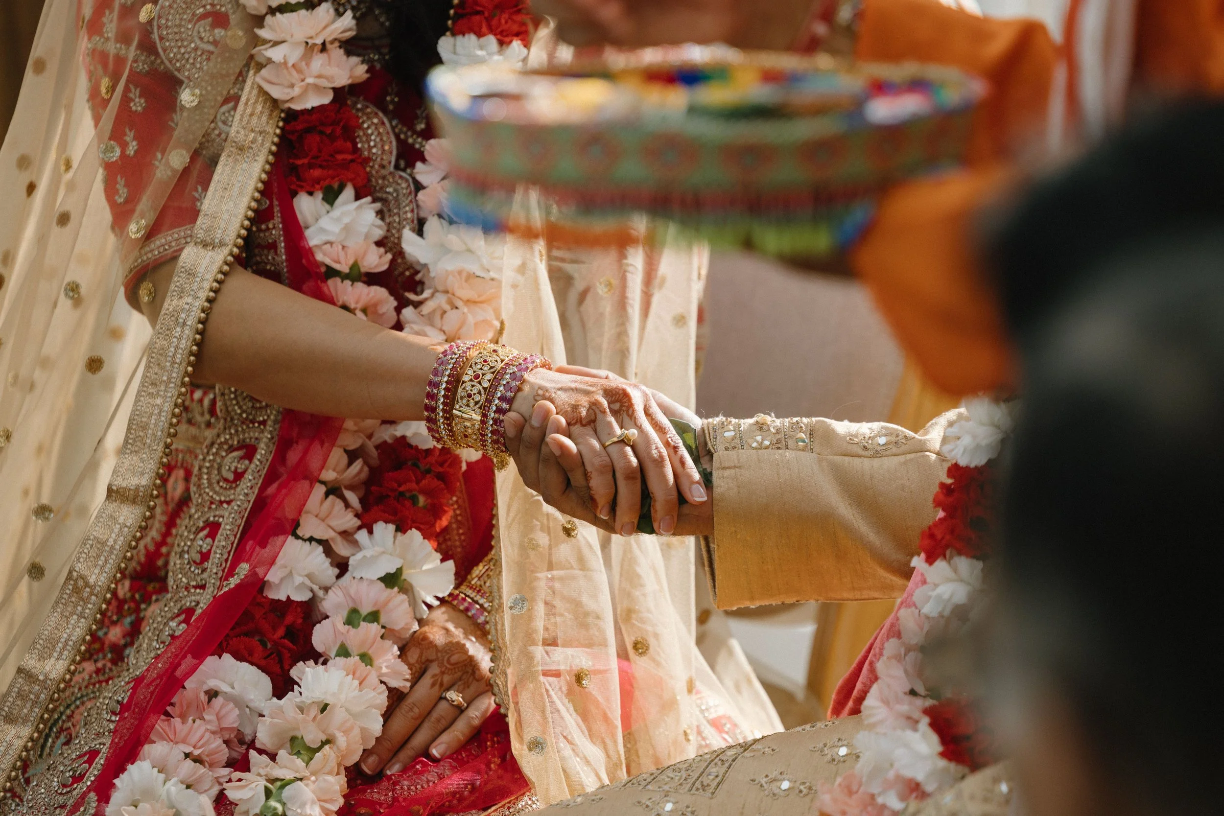 Close-up of a couple holding hands during a traditional wedding ceremony, with the bride wearing ornate jewelry and a richly decorated red and gold sari, surrounded by flower arrangements.