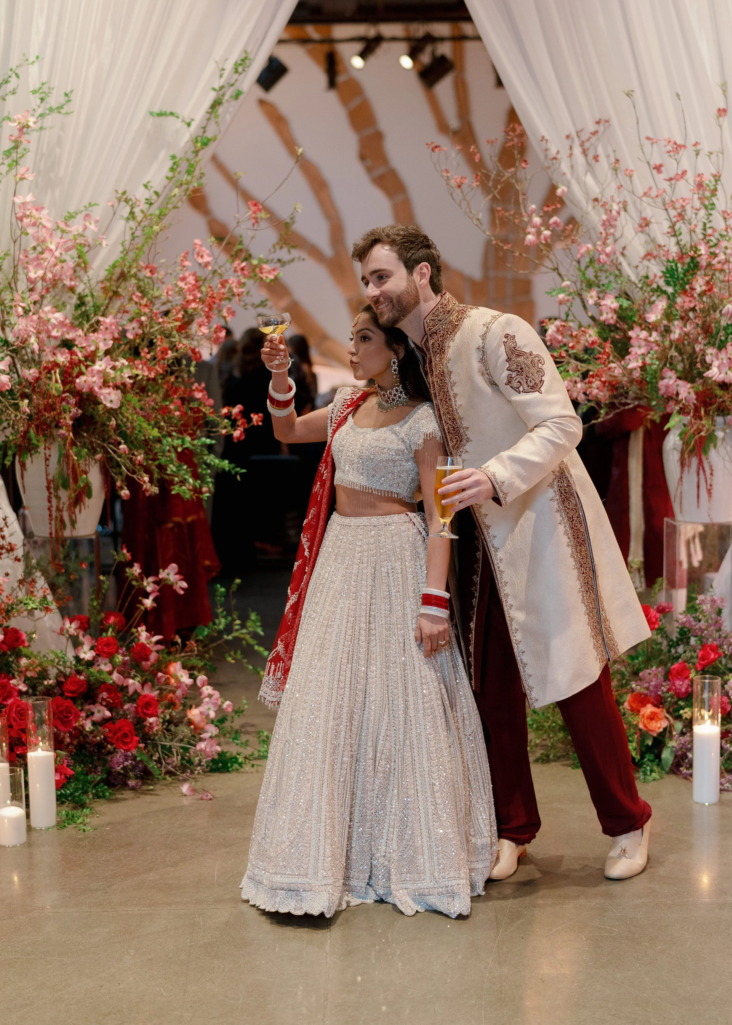 A couple in traditional Indian wedding attire celebrating together at their wedding reception. The woman is holding a champagne glass, and the man is holding a glass of champagne. They are surrounded by pink and red floral arrangements and candles.
