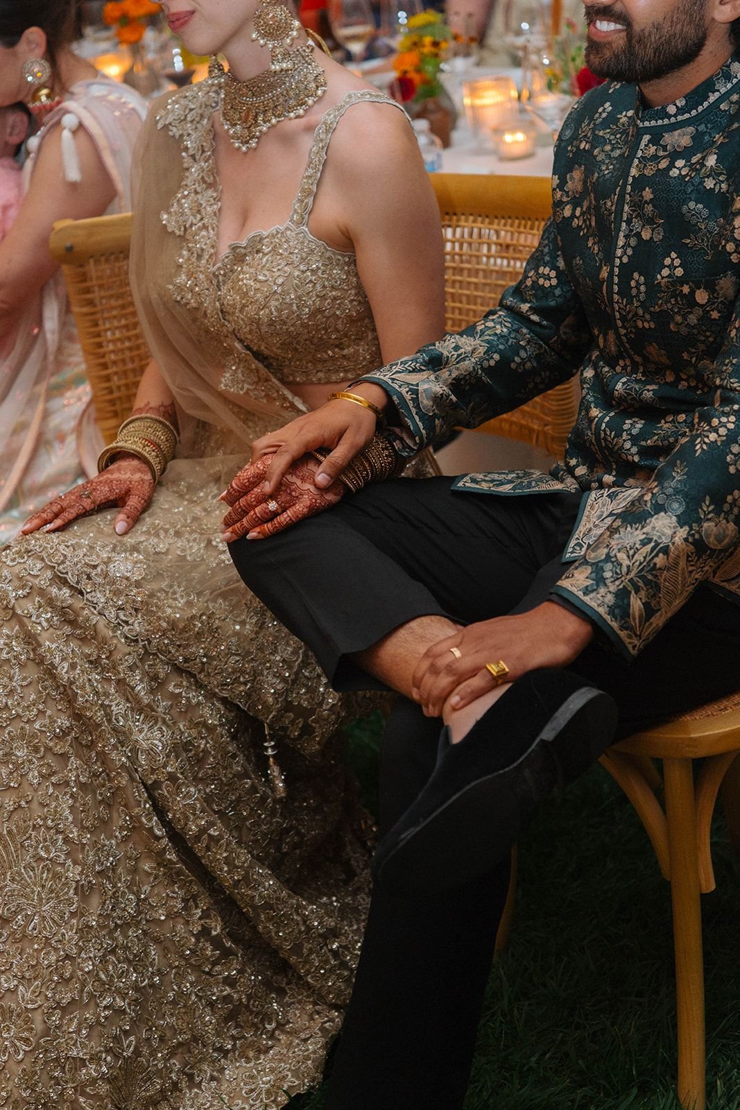 A bride and groom sitting holding hands at a wedding with traditional Indian attire and jewelry, and wedding decorations in the background.