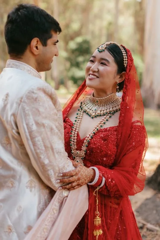 A bride and groom in traditional Indian wedding attire smiling and holding hands outdoors during the day.