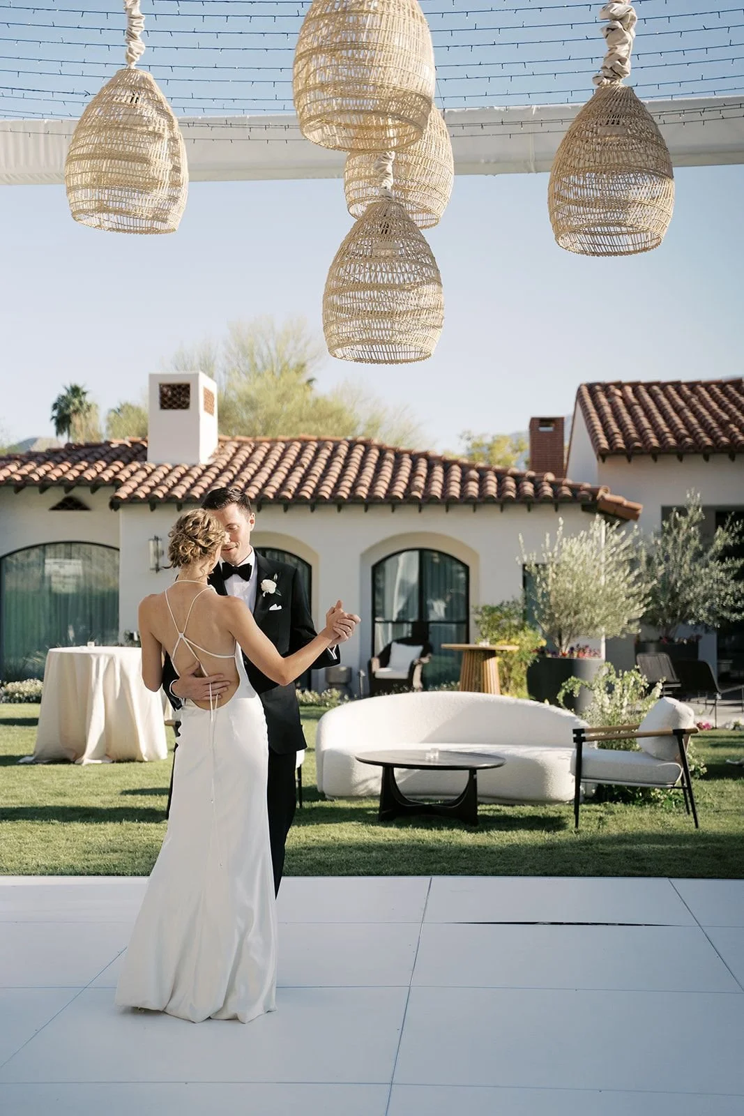 A bride and groom dancing outdoors at a wedding reception with a modern house and string lights above.