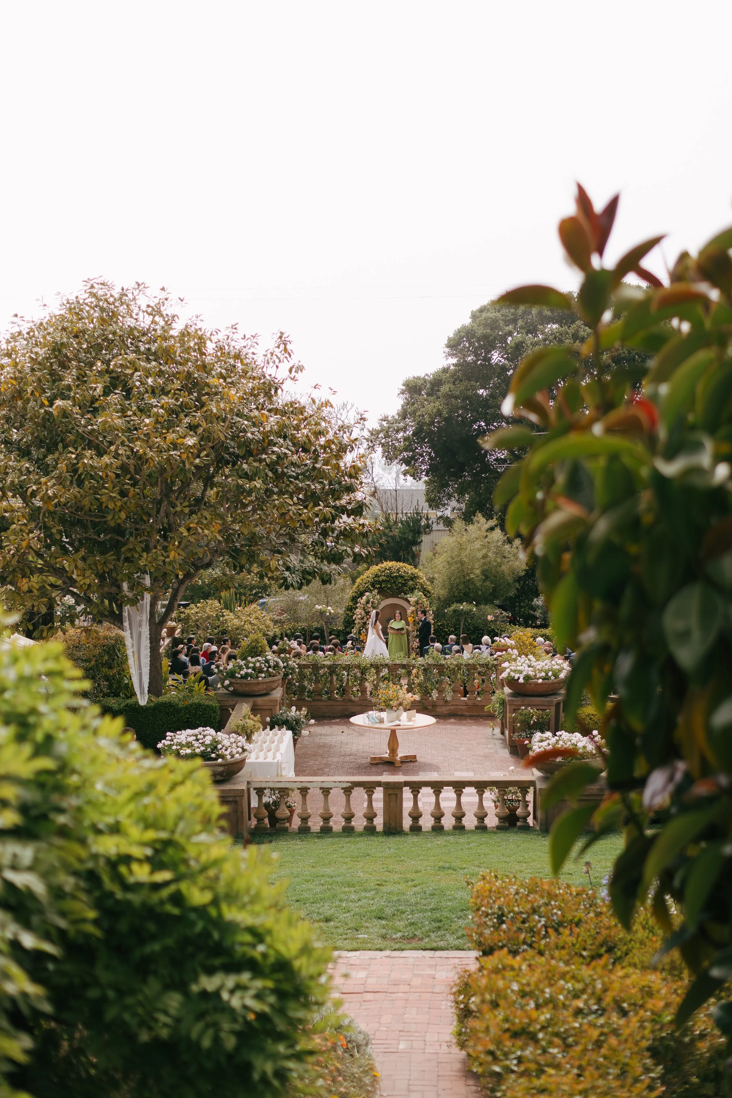 An outdoor wedding ceremony taking place in a lush garden, surrounded by trees and floral arrangements, with guests seated on chairs and a couple standing before an officiant.