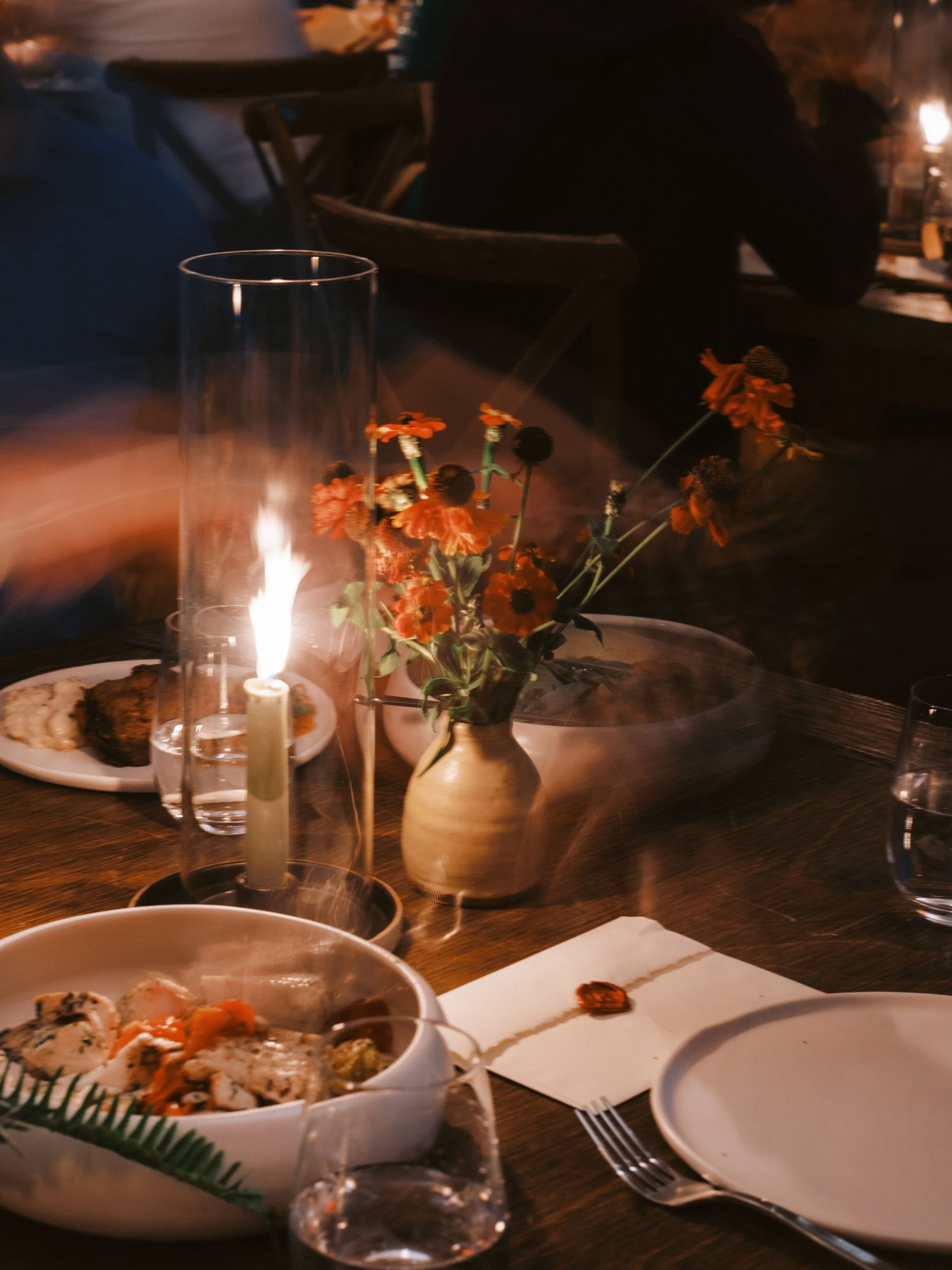 Close-up of a dining table with plates of food, a glass candle holder, a vase with orange flowers, and a lit candle in the background, with a person wearing dark clothing leaning over the table.