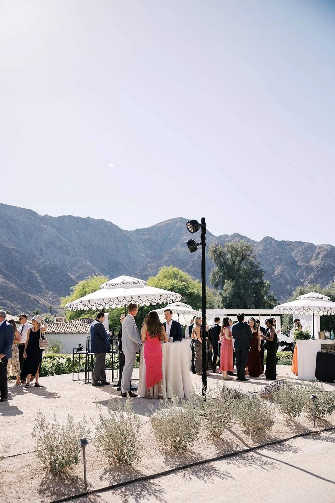 People attending an outdoor event at a venue with mountain views, white umbrellas, and large lighting fixtures, with some individuals engaged in conversations around high tables.