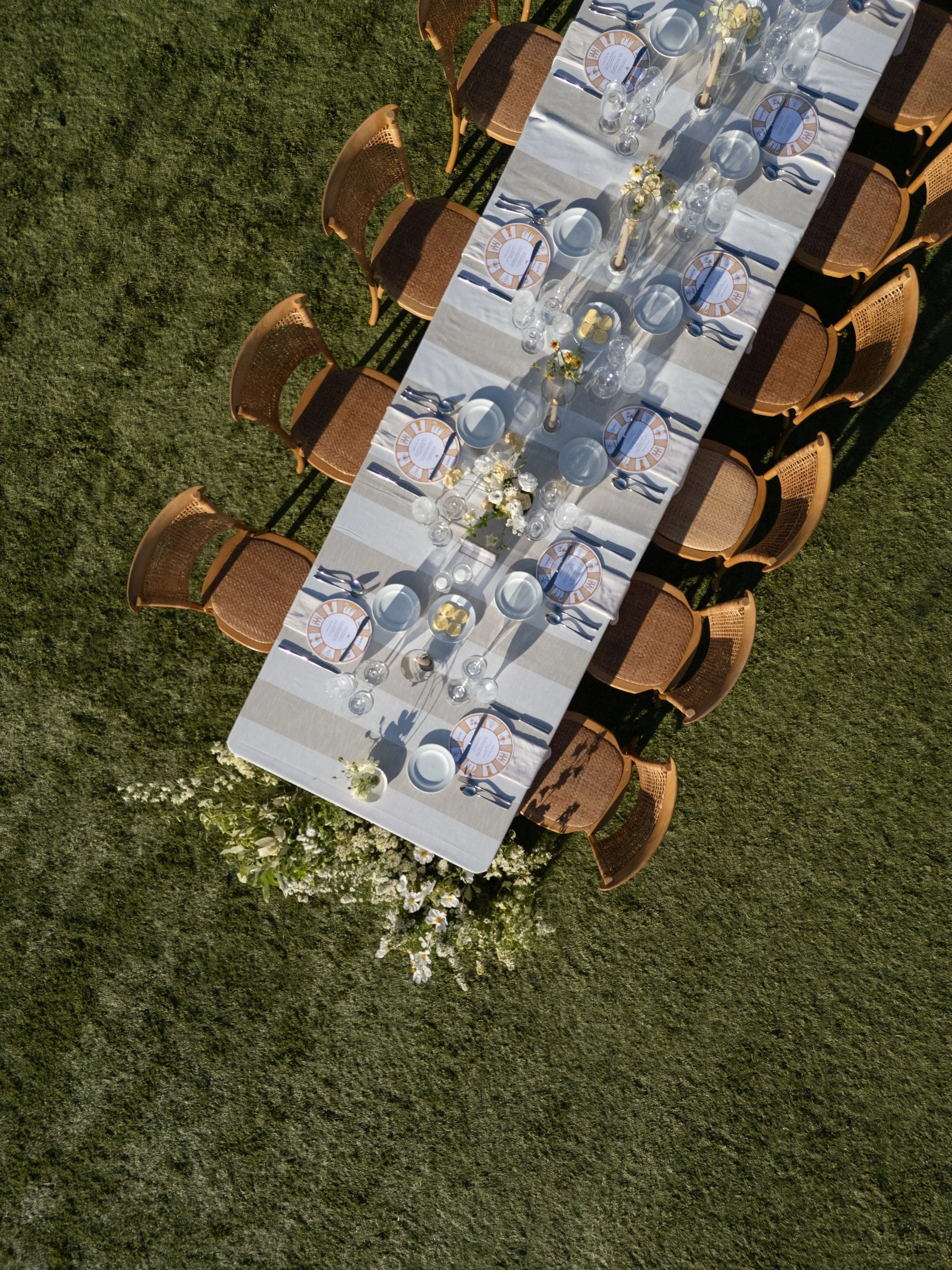 An outdoor banquet table set for a meal on a grassy area, with wooden chairs, a white tablecloth, plates, glasses, utensils, small flower vases, and decorative chargers.