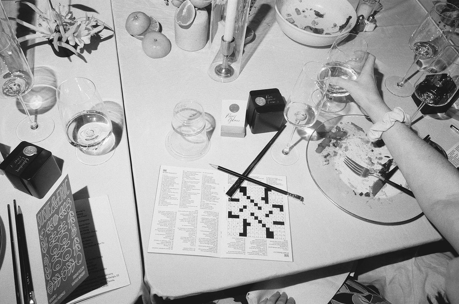 A dinner table with empty wine glasses, a pizza crust on a plate, a hand holding a wine glass, a crossword puzzle, and various items like a menu, a candle holder, and some fruit in the background.