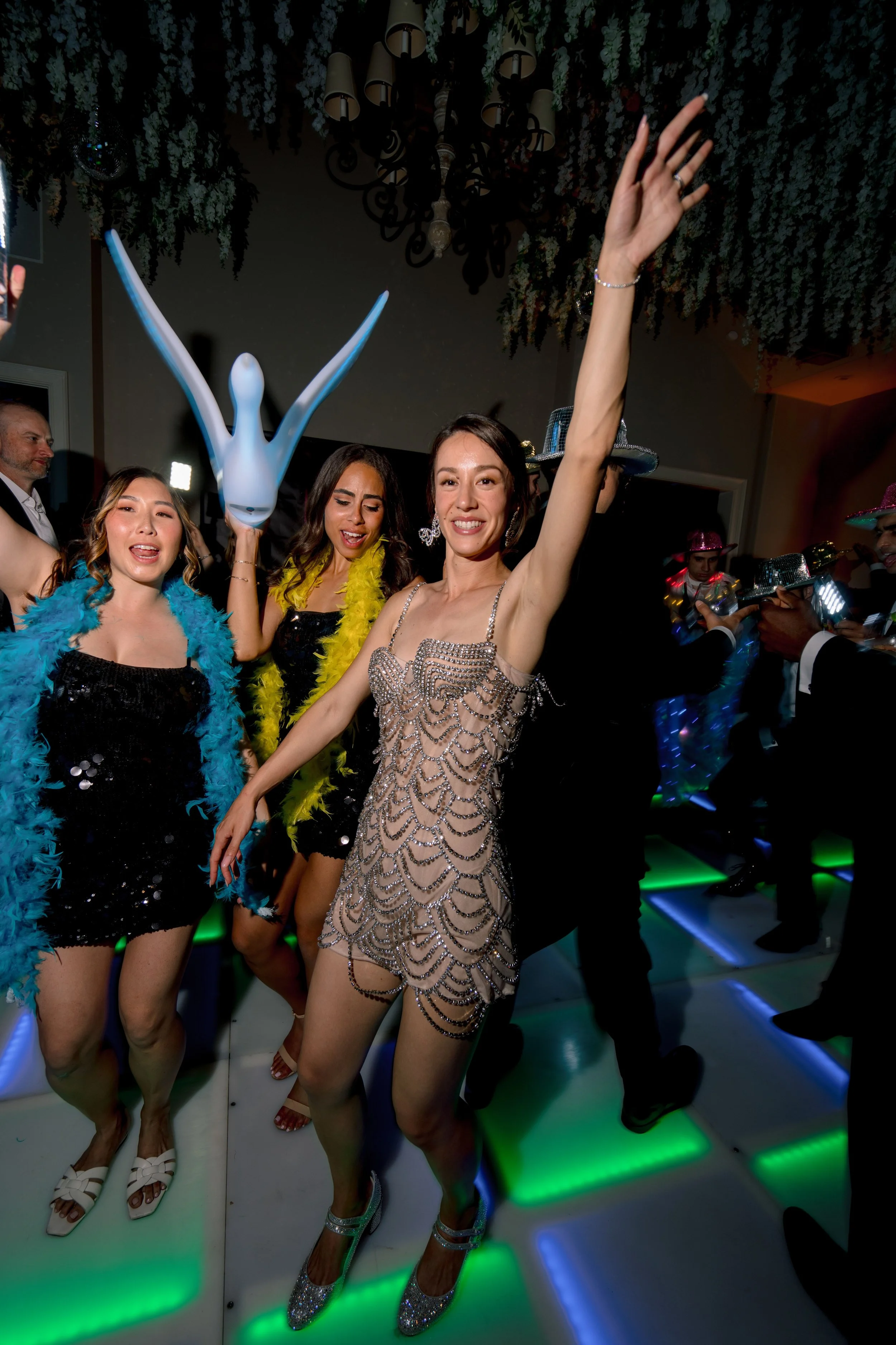 Three women dancing at a party, one in a beaded dress, another in a black dress with a yellow feather boa, and the third in a black dress with a turquoise feather boa, with party hats and colorful lighting.