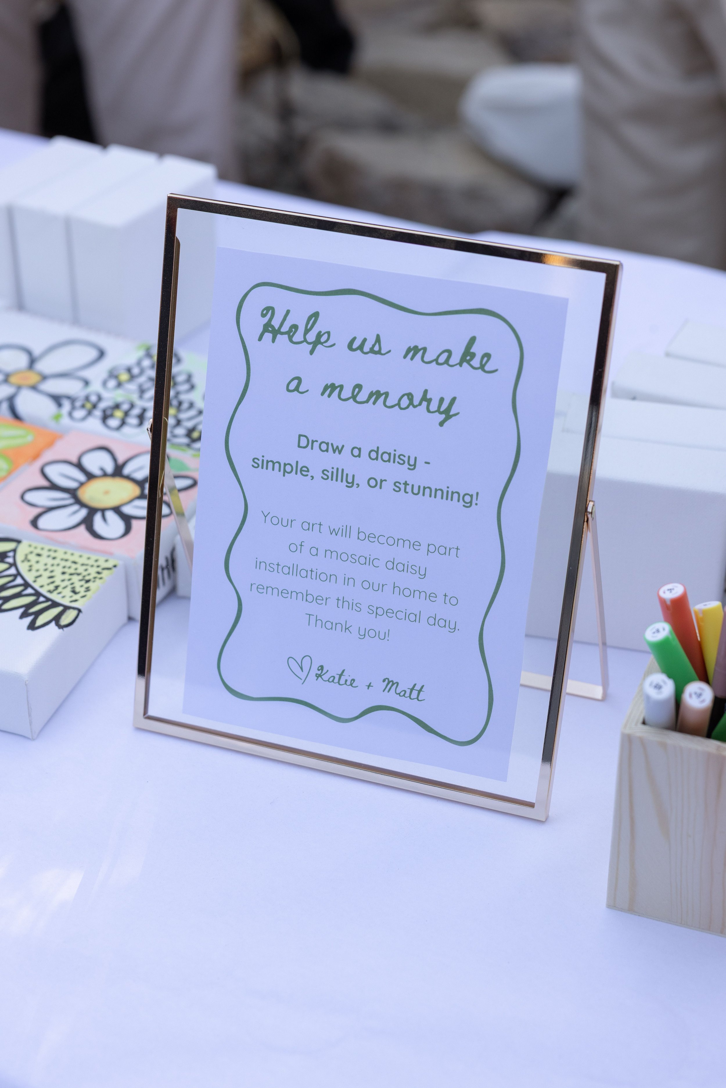 Sign that reads 'Help us make a memory. Draw a daisy - simple, silly, or stunning!', with a background showing some artwork and markers on a table, possibly at a wedding or event.