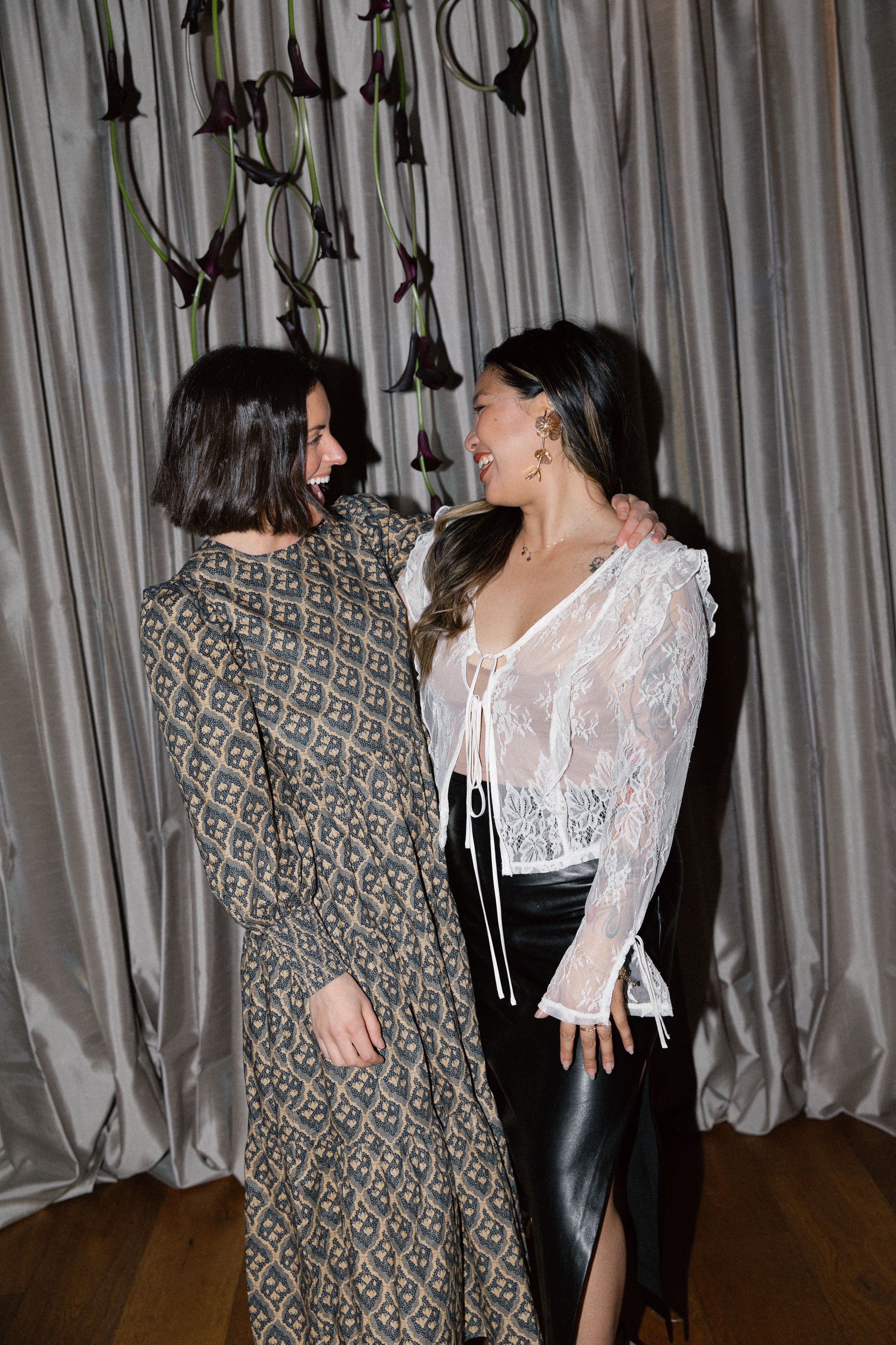Two women are smiling and looking at each other while standing close together in front of a grey curtain with purple and black hanging flowers. One woman has short dark hair and is wearing a patterned dress, while the other has long dark hair and is 