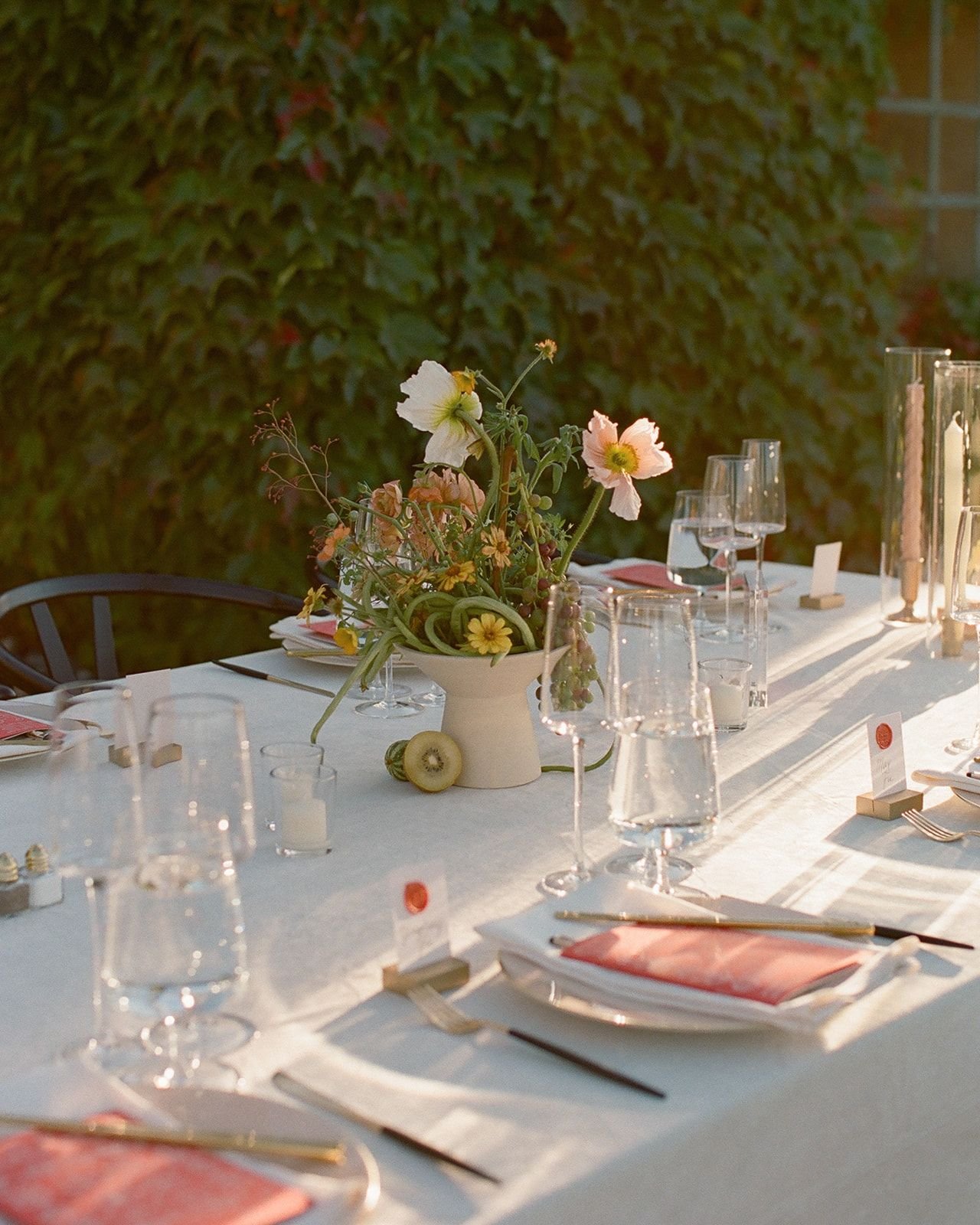 A long dining table set for an outdoor dinner, decorated with a white tablecloth, glassware, pink napkins, and a centerpiece of pastel-colored flowers in a white vase, with candle holders and a lush green hedge in the background.
