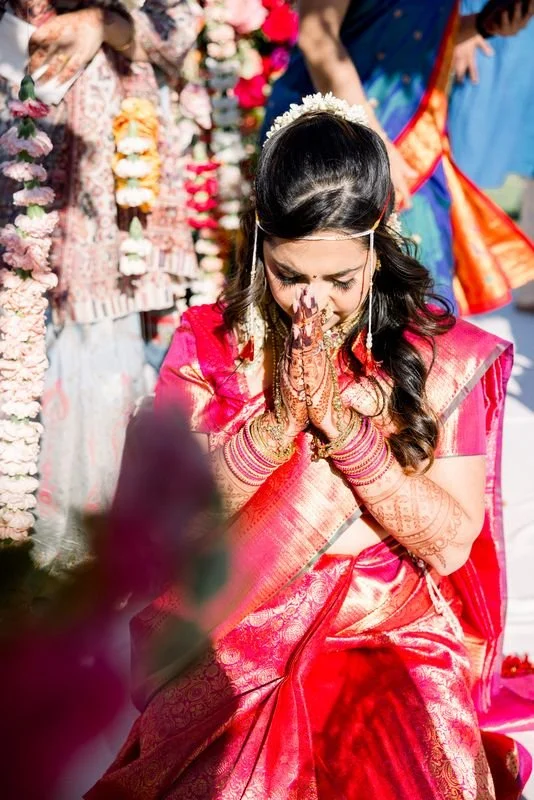 A woman dressed in a pink and gold traditional Indian saree, bowing with hands pressed together in prayer, adorned with jewelry and henna designs, during a religious or cultural ceremony.